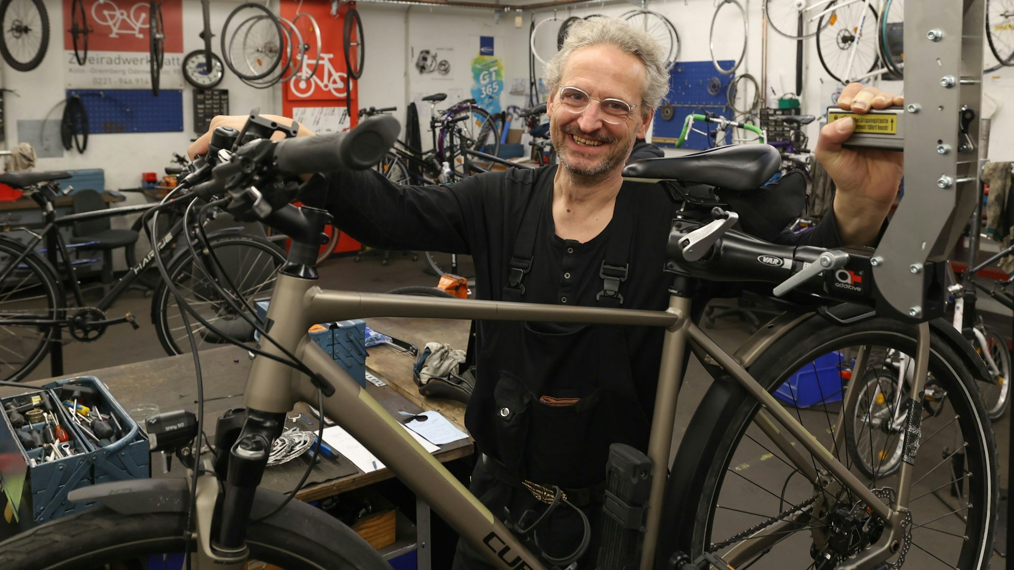 Ein Mann im schwarzen Overall, mit Brille und grauem Haar steht hinter einem Fahrrad in einer Werkstatt.