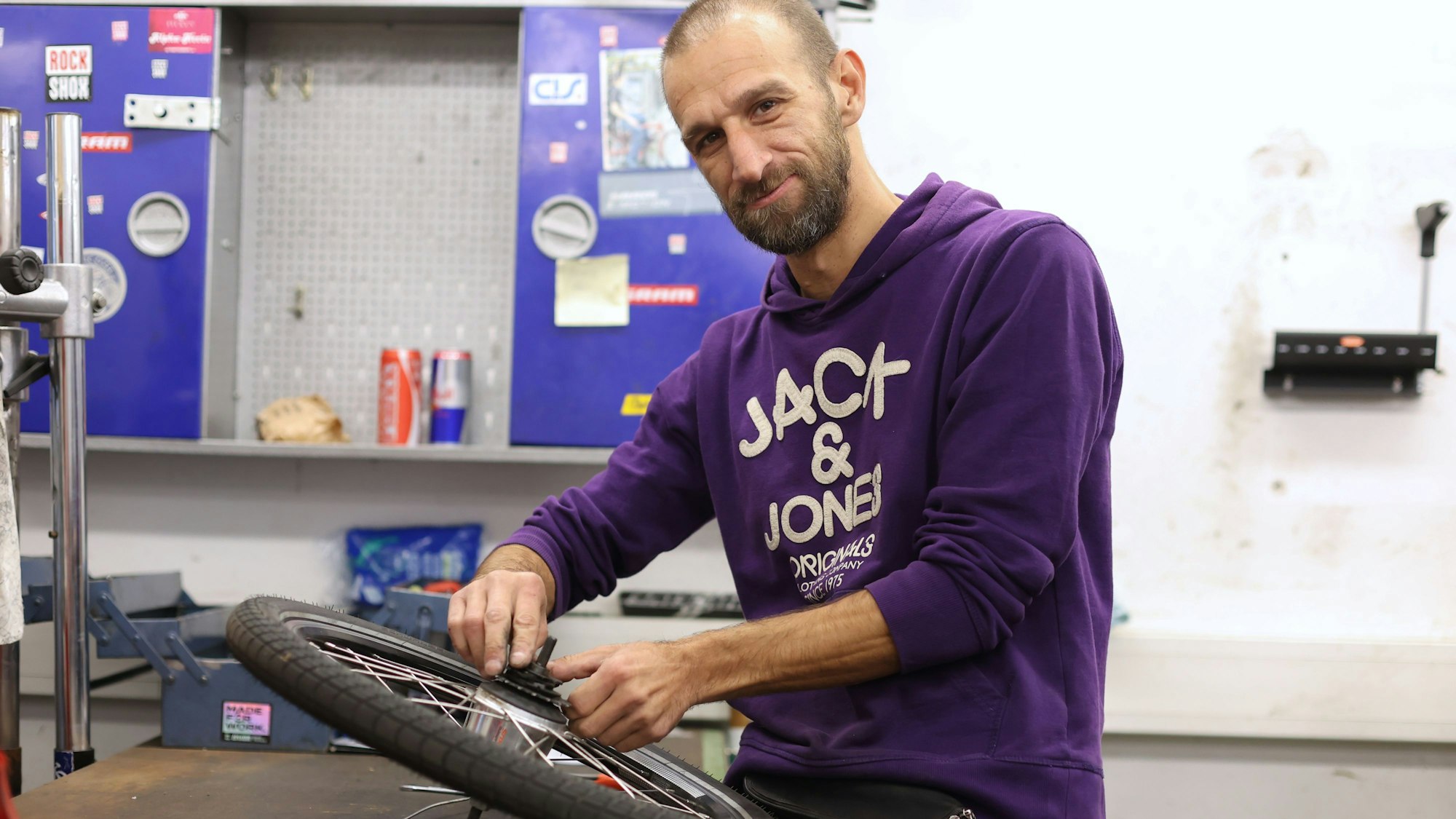Ein Mann mit Bart und lila Hoodie arbeitet an einem Fahrradreifen, das vor ihm auf einer Werkbank liegt.