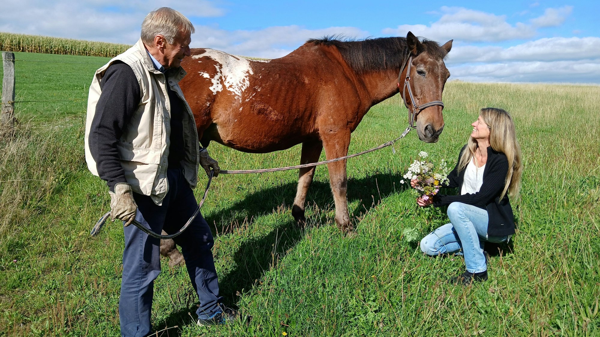 Eine Frau hält einen Blumenstrauß in der Hand und kniet auf einer Wiese vor einem braunen Pferd. Ein Mann hält das Pferd an einem Strick.