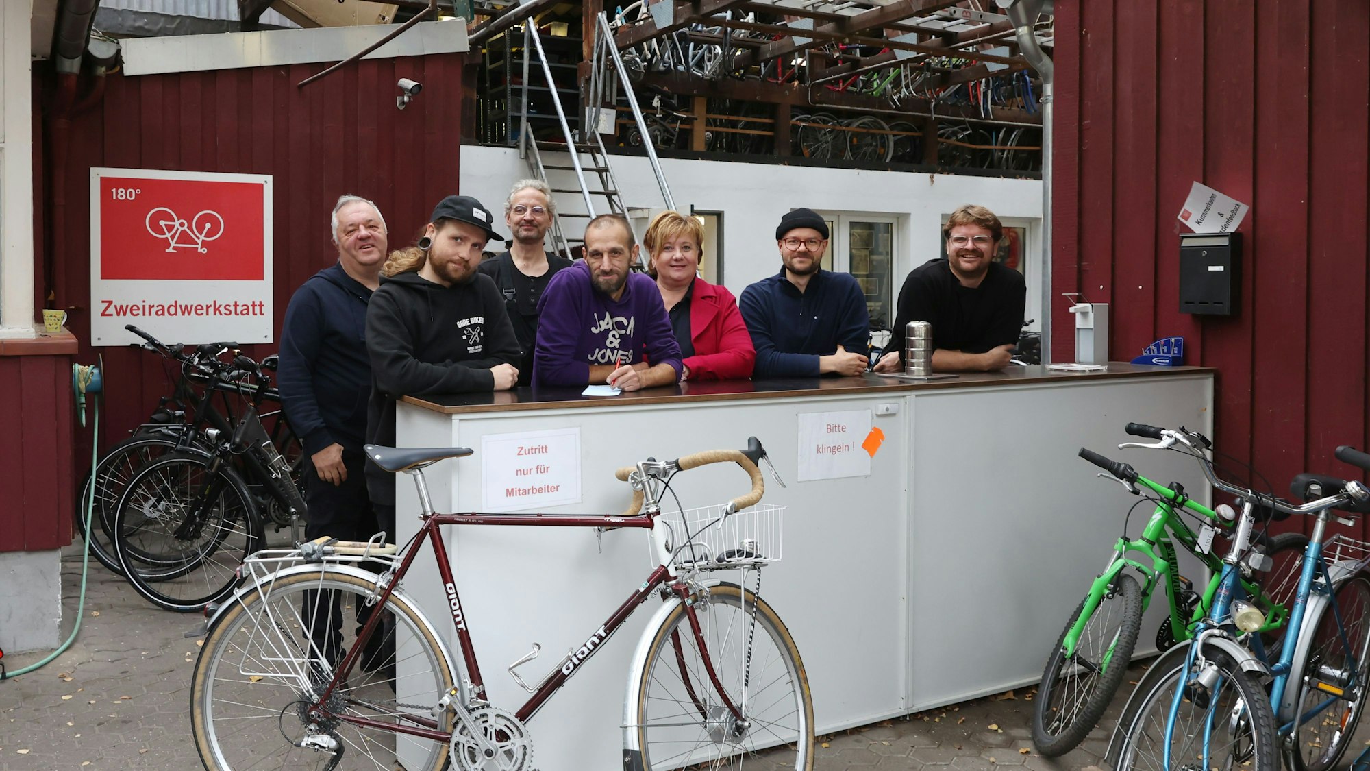 Mehrere Menschen stehen hinter einer Art Theke, daran lehnt ein Fahrrad, weitere Kinderräder stehen am Rand.