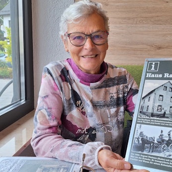 Renate Krumpen sitzt an einem Tisch und hält eine Tafel in der Hand, die alte Ansichten eines Hauses zeigt.