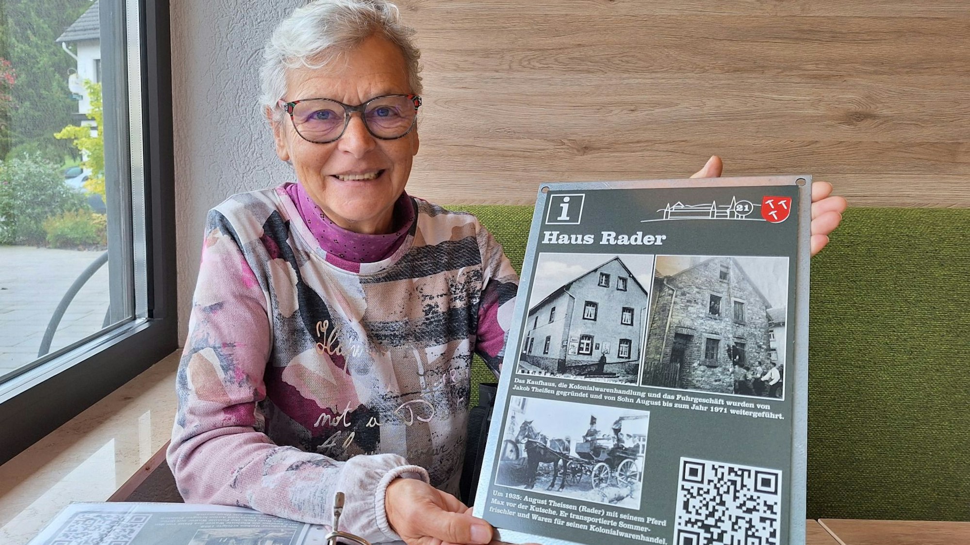 Renate Krumpen sitzt an einem Tisch und hält eine Tafel in der Hand, die alte Ansichten eines Hauses zeigt.