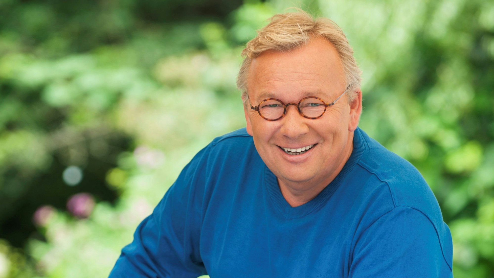 Bernd Stelter trägt einen blauen Pulli, eine runde Brille und lächelt. Er schiebt ein Fahrrad. Im Hintergrund viel Grün.