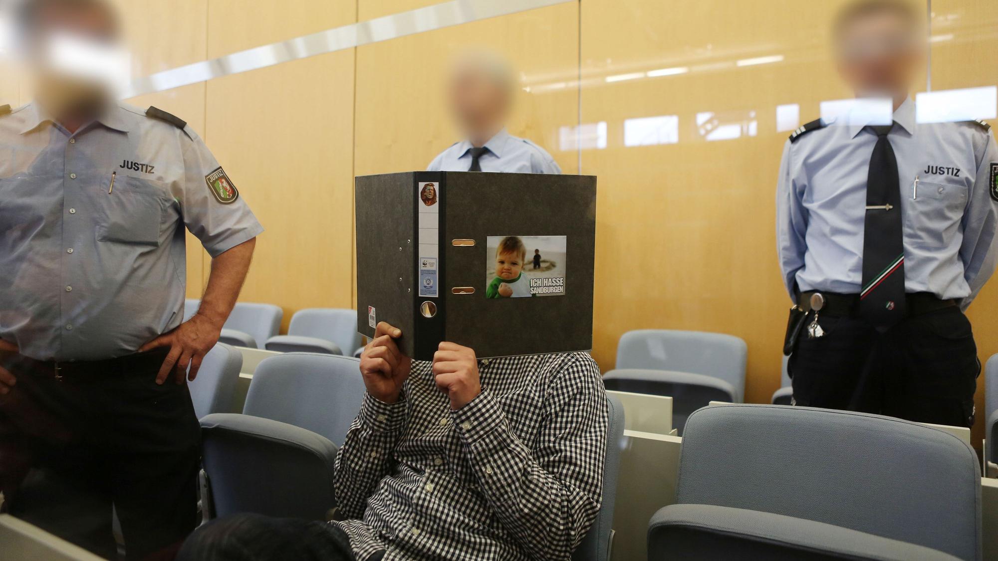 Der Angeklagte Frank S. sitzt am 15.04.2016 in Düsseldorf (Nordrhein-Westfalen) umringt von Jutizbeamten im Verhandlungssaal und hält sich einen Aktendeckel vor das Gesicht. Der Prozess vor dem Oberlandesgericht um das Attentat auf Henriette Reker, einen Tag vor ihrer Wahl zur Kölner Oberbürgermeisterin, hat begonnen. Foto: Oliver Berg/dpa +++(c) dpa - Bildfunk+++