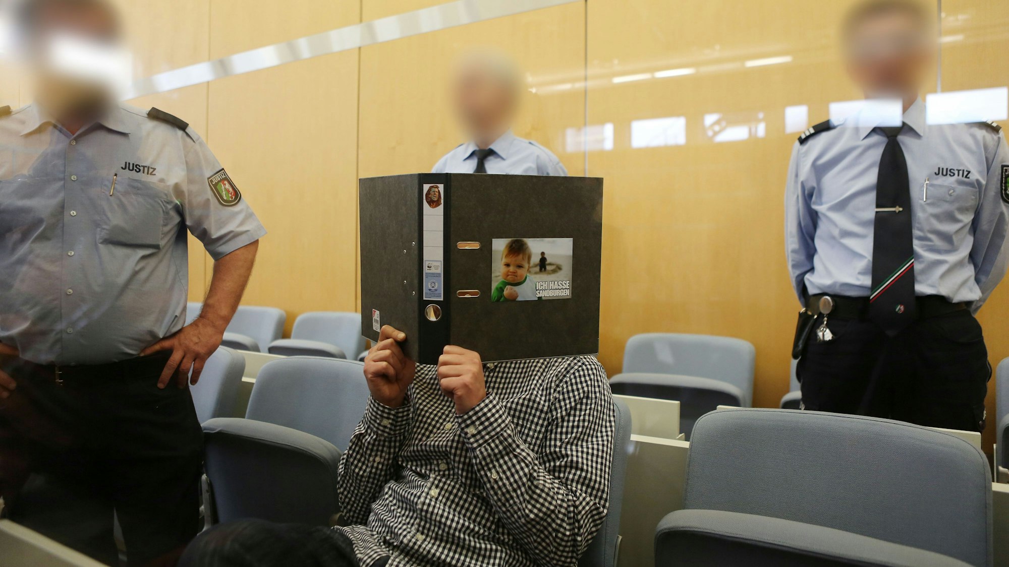 Der Angeklagte Frank S. sitzt am 15.04.2016 in Düsseldorf (Nordrhein-Westfalen) umringt von Jutizbeamten im Verhandlungssaal und hält sich einen Aktendeckel vor das Gesicht. Der Prozess vor dem Oberlandesgericht um das Attentat auf Henriette Reker, einen Tag vor ihrer Wahl zur Kölner Oberbürgermeisterin, hat begonnen. Foto: Oliver Berg/dpa +++(c) dpa - Bildfunk+++