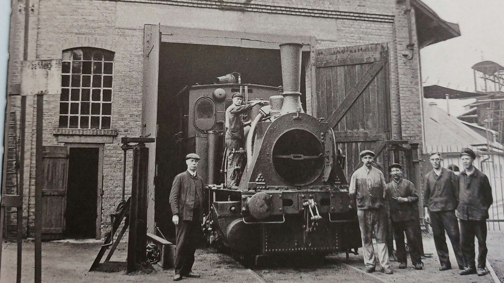 Schwarz-weiß-Foto einer Lokomotive auf einem Betriebsgelände, links und rechts stehen Arbeiter.