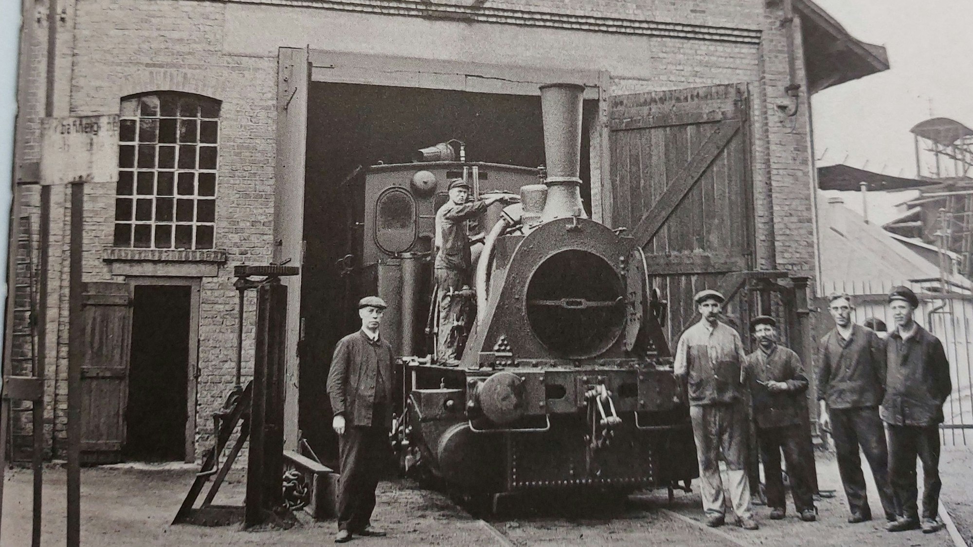 Schwarz-weiß-Foto einer Lokomotive auf einem Betriebsgelände, links und rechts stehen Arbeiter.