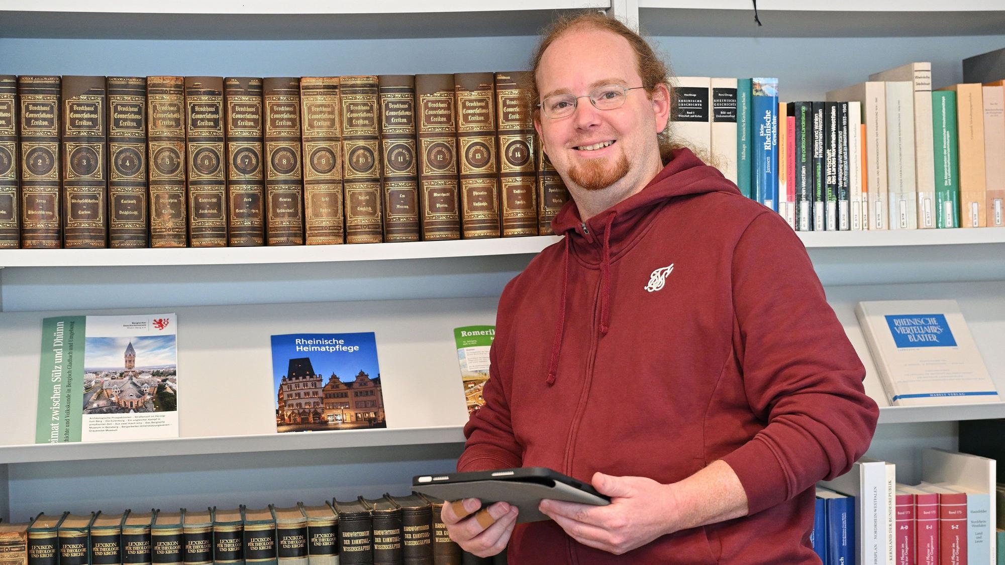 Ein Mann in rotem Kapuzenpulli, mit rotem Haar und rotem Bart, steht vor einem Bücherregal mit alten Nachschlagewerken.