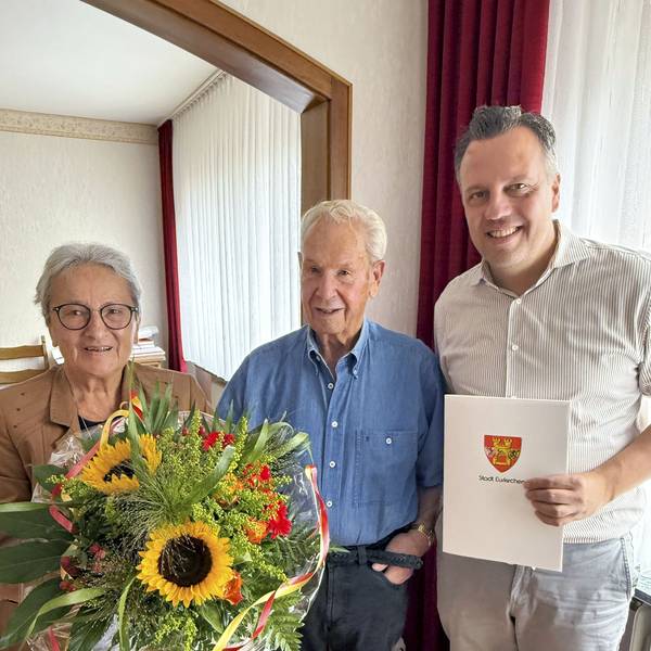 Anneliese Büschkes hält einen Blumenstrauß, Bürgermeister Sacha Reichelt überreicht Hans-Dieter Büschkes eine Urkunde, auf der das Wappen der Stadt Euskirchen zu sehen ist.