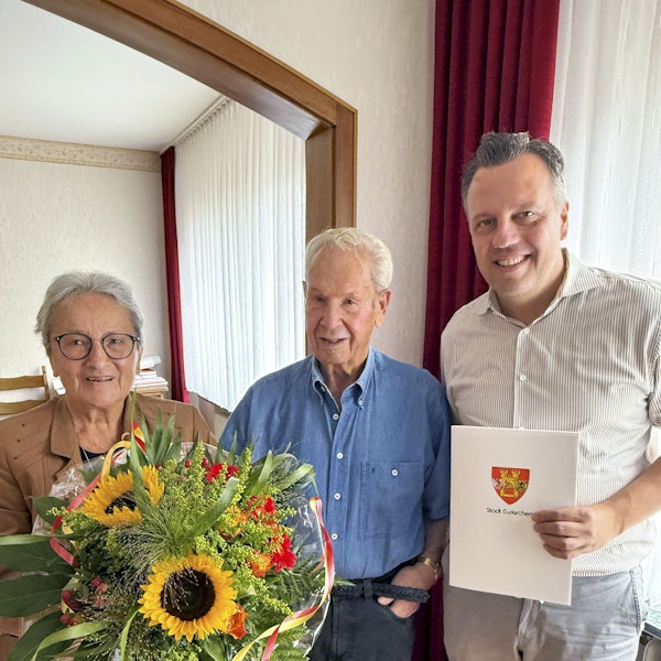 Anneliese Büschkes hält einen Blumenstrauß, Bürgermeister Sacha Reichelt überreicht Hans-Dieter Büschkes eine Urkunde, auf der das Wappen der Stadt Euskirchen zu sehen ist.