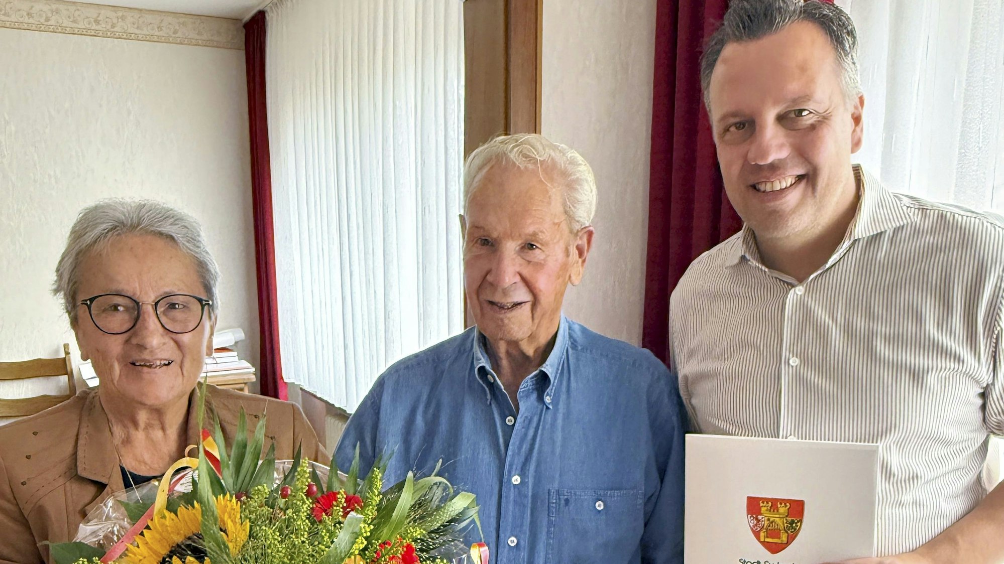 Anneliese Büschkes hält einen Blumenstrauß, Bürgermeister Sacha Reichelt überreicht Hans-Dieter Büschkes eine Urkunde, auf der das Wappen der Stadt Euskirchen zu sehen ist.