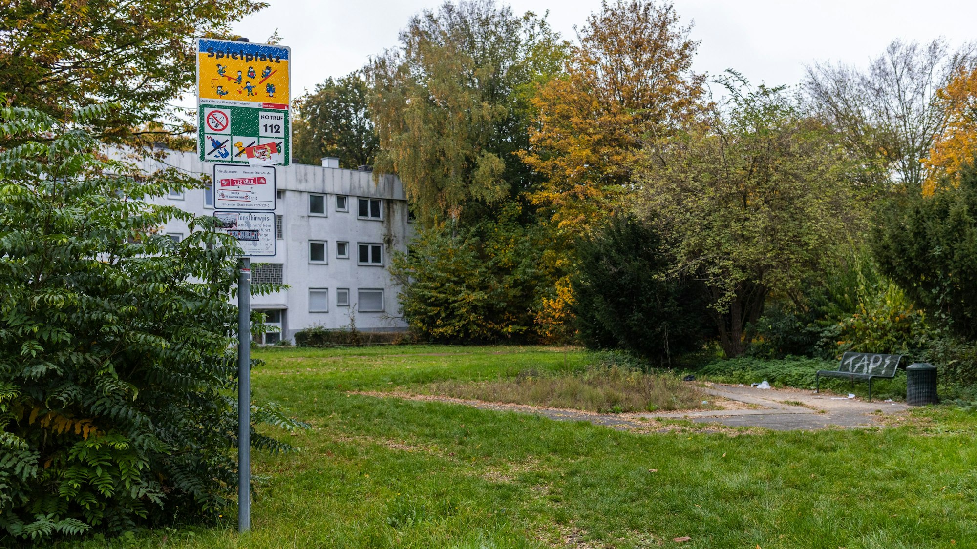 Eine Fläche mit Gras und einer Bank. Davor steht ein Spielplatzschild.