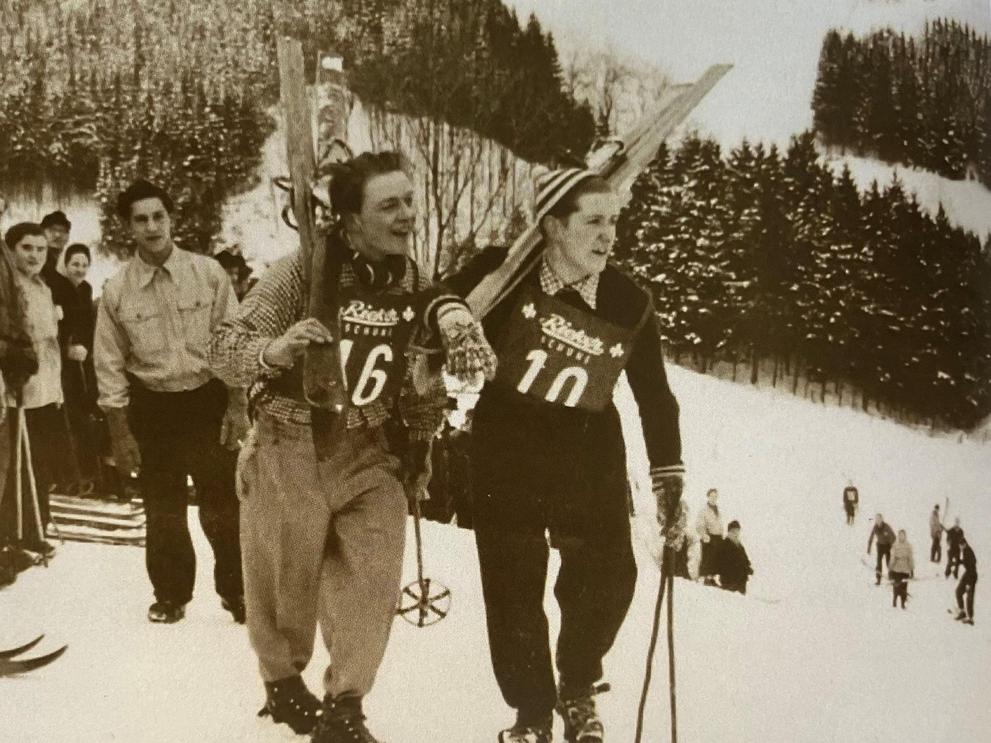 Ein Schwarz-Weiß-Foto: Zwei Skiläufer tragen ihre Ski einen Berg hoch.