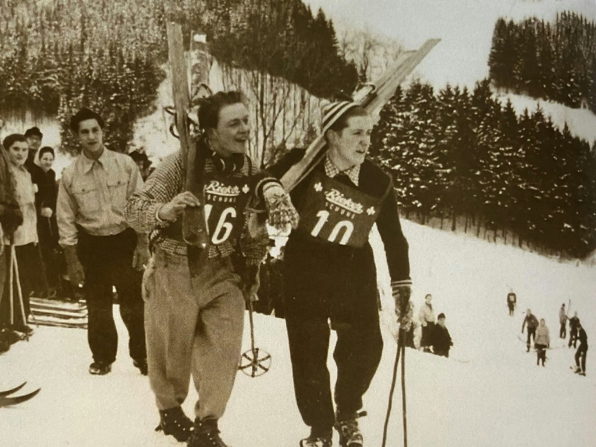 Ein Schwarz-Weiß-Foto: Zwei Skiläufer tragen ihre Ski einen Berg hoch.