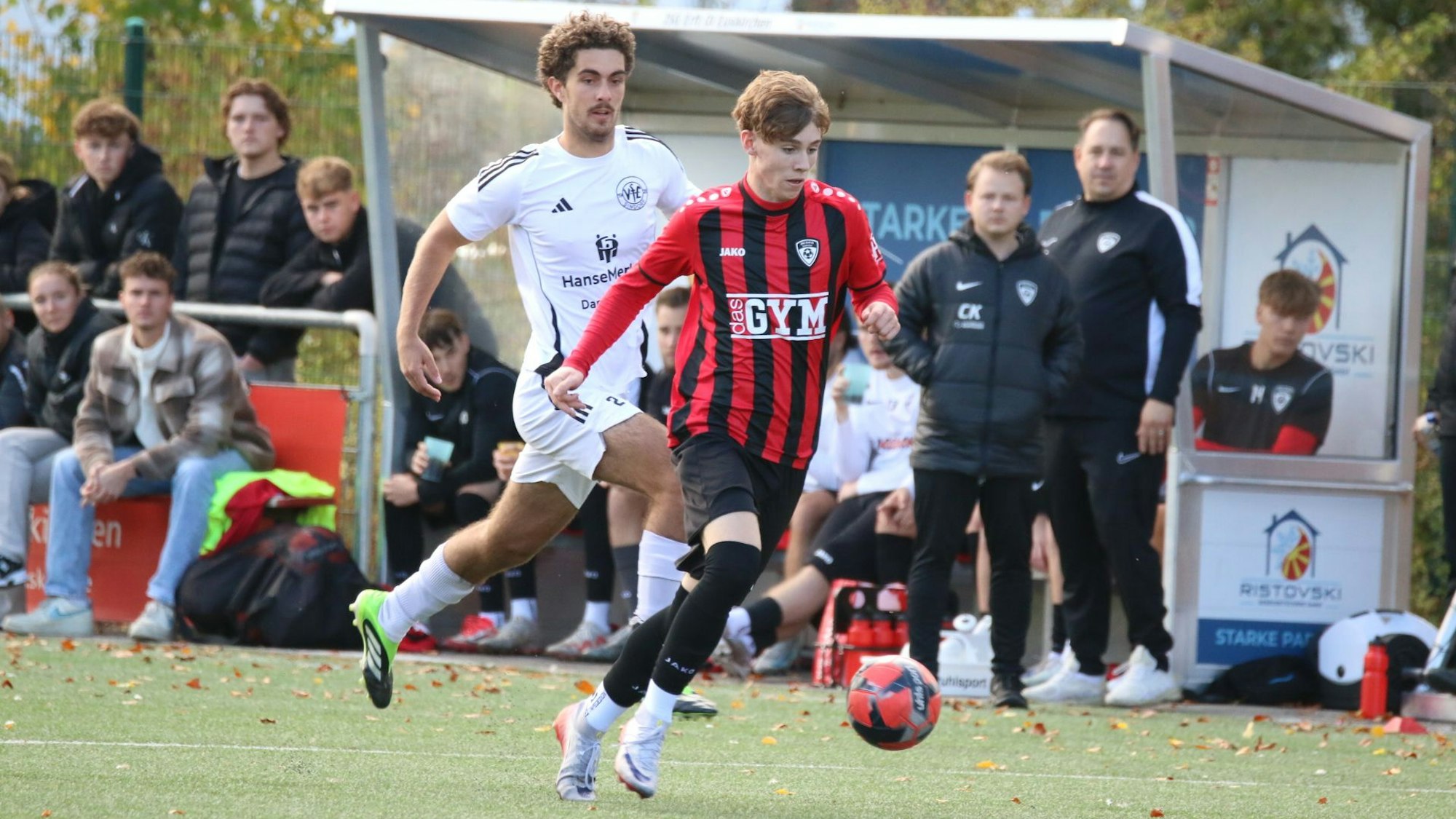 Aaron Mertens läuft mit dem Ball, ein Gegenspieler verfolgt ihn. Im Hintergrund gucken Trainer und Betreuer der JSG Erft 01 zu.