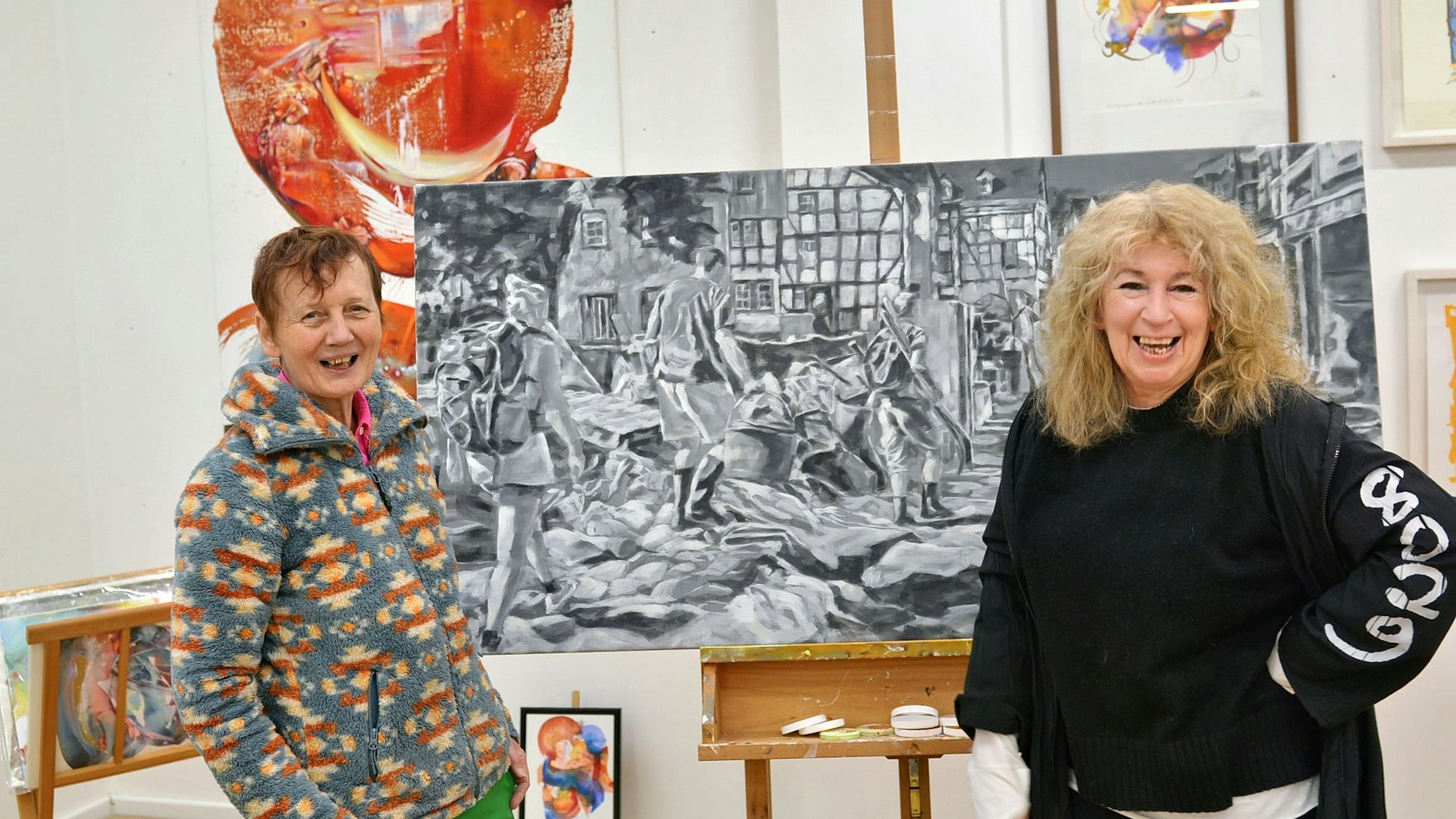 Gaby Kutz und Maf Räderscheidt blicken lachend zur Kamera. Im Hintergrund stehen und hängen Gemälde beider Frauen.