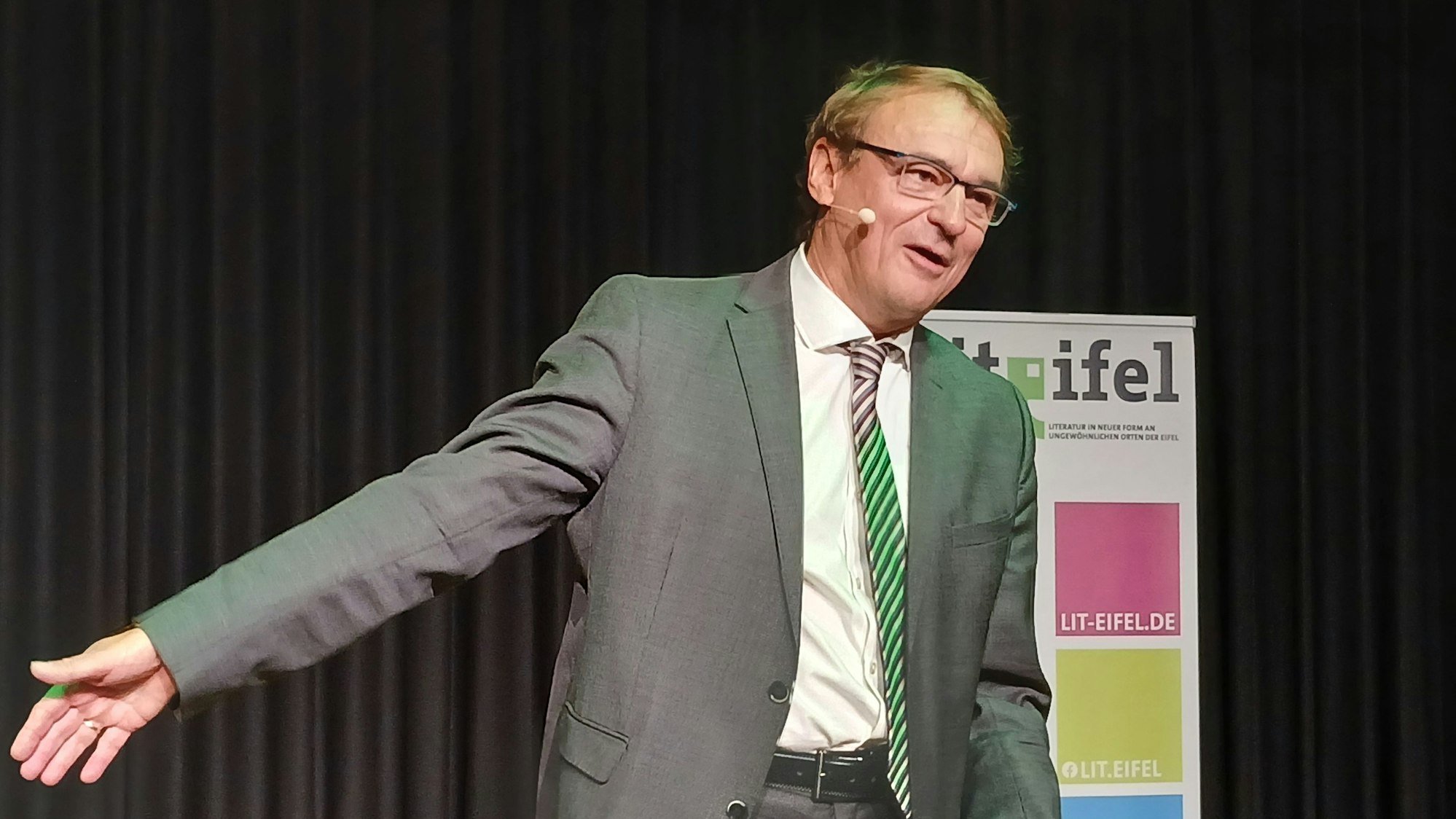 Andreas Englisch steht im grauen Anzug mit bunter Krawatte auf der Bühne und gestikuliert, während er redet.