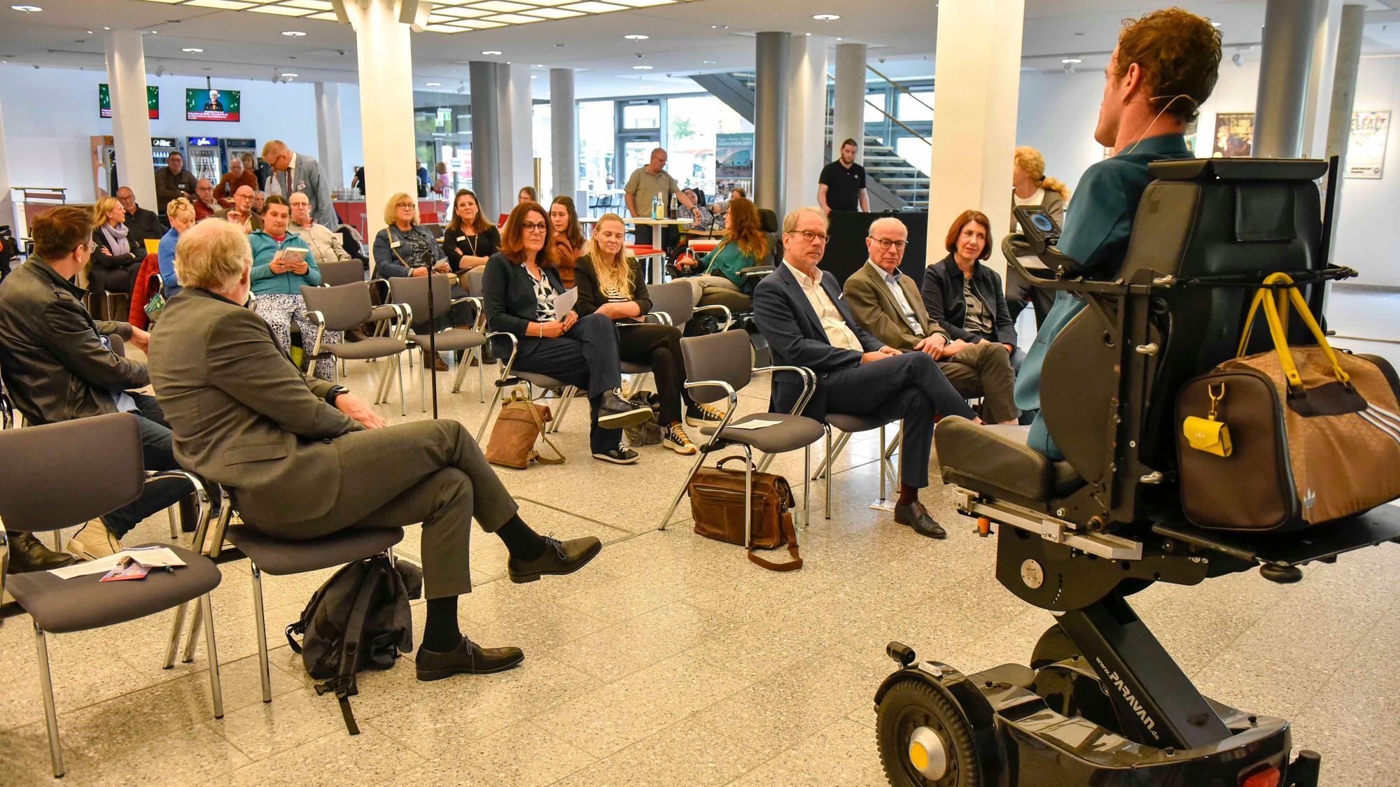 Blick in einen Saal während eines Vortrags. Rechts im Bild, in einem Rollstuhl, der Vortragsredner Janis McDavid, der ohne Arme und Beine zur Welt kam.