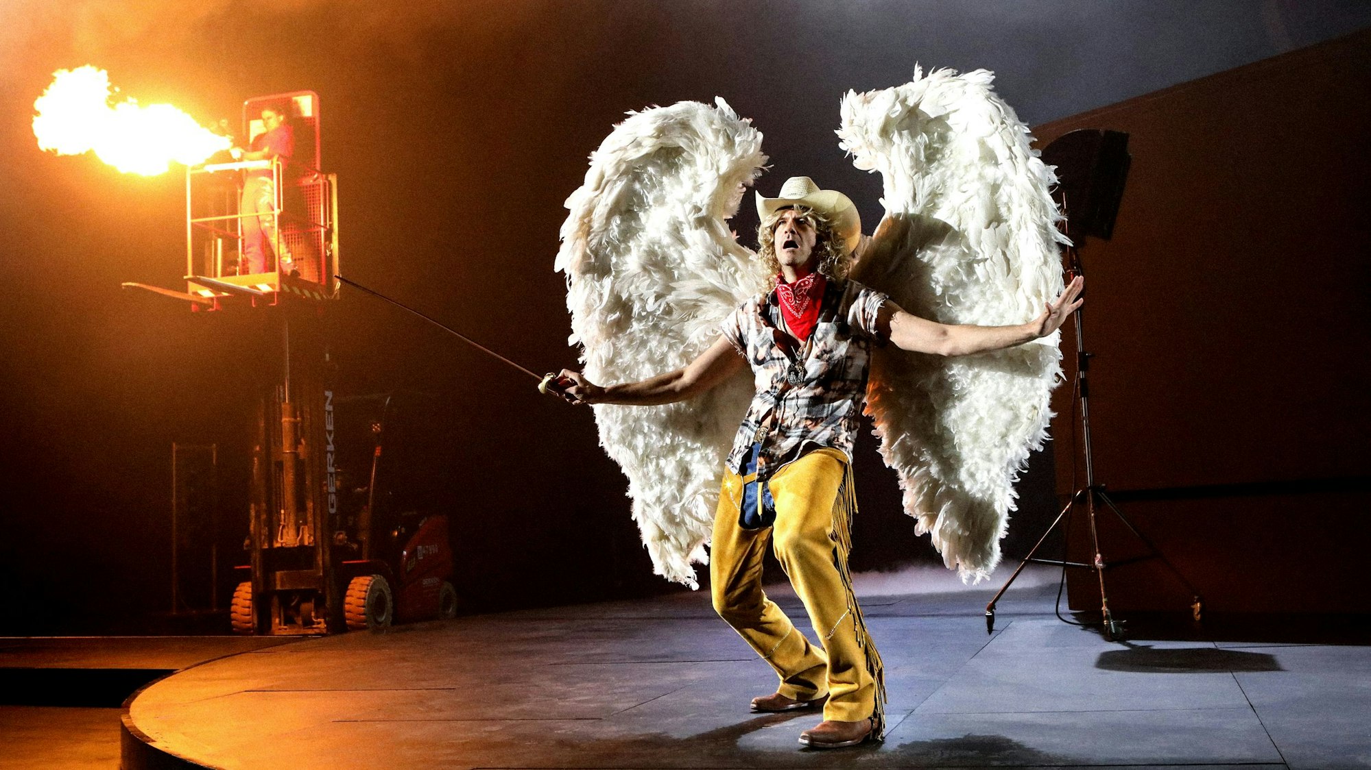 Lavinia Nowak steht auf einem Gabelstapler und betätigt einen Flammenwerfer, Frank Genser trägt große Engelsflügel und Cowboyhut