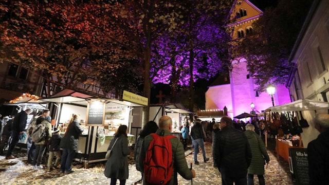 Menschen an mehreren Marktbuden, im Hintergrund eine von Scheinwerfern angestrahlte Kirche.