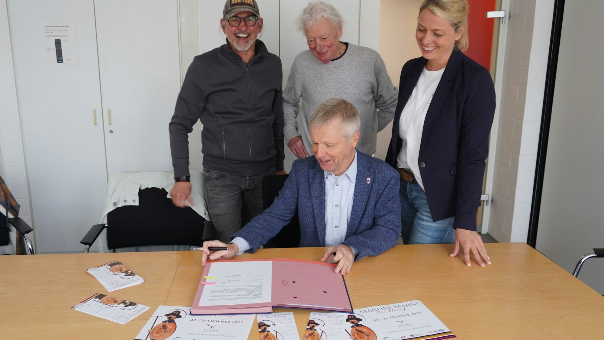 Drei Männer und eine Frau. Ein Mann sitzt an einem Tisch vor einer Dokumentenmappe.