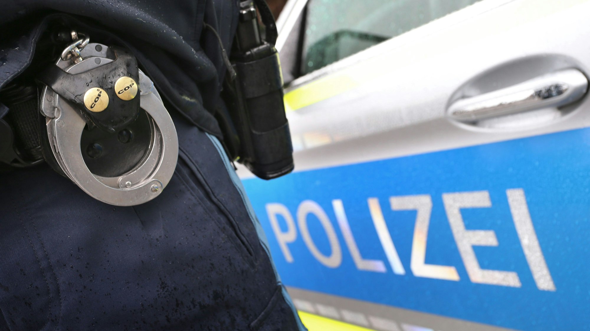 Ein Polizist mit Handschellen und einer Pistole am Gürtel steht vor einem Streifenwagen.
