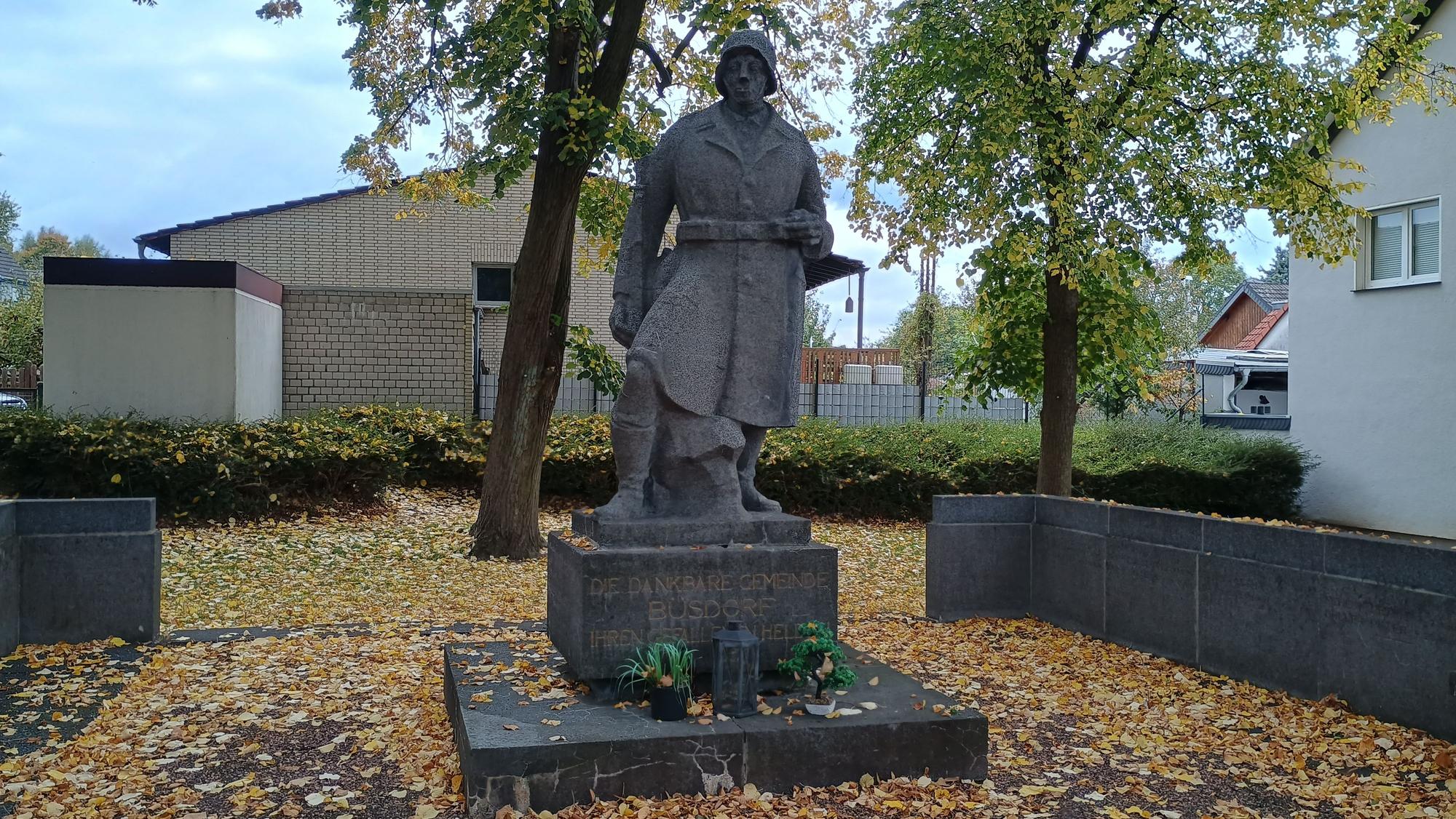 Zu sehen ist ein Steinsoldat mit Helm und Uniform. Auf seinem Podest steht: „Die dankbare Gemeinde Büsdorf ihren gefallenen Helden.“