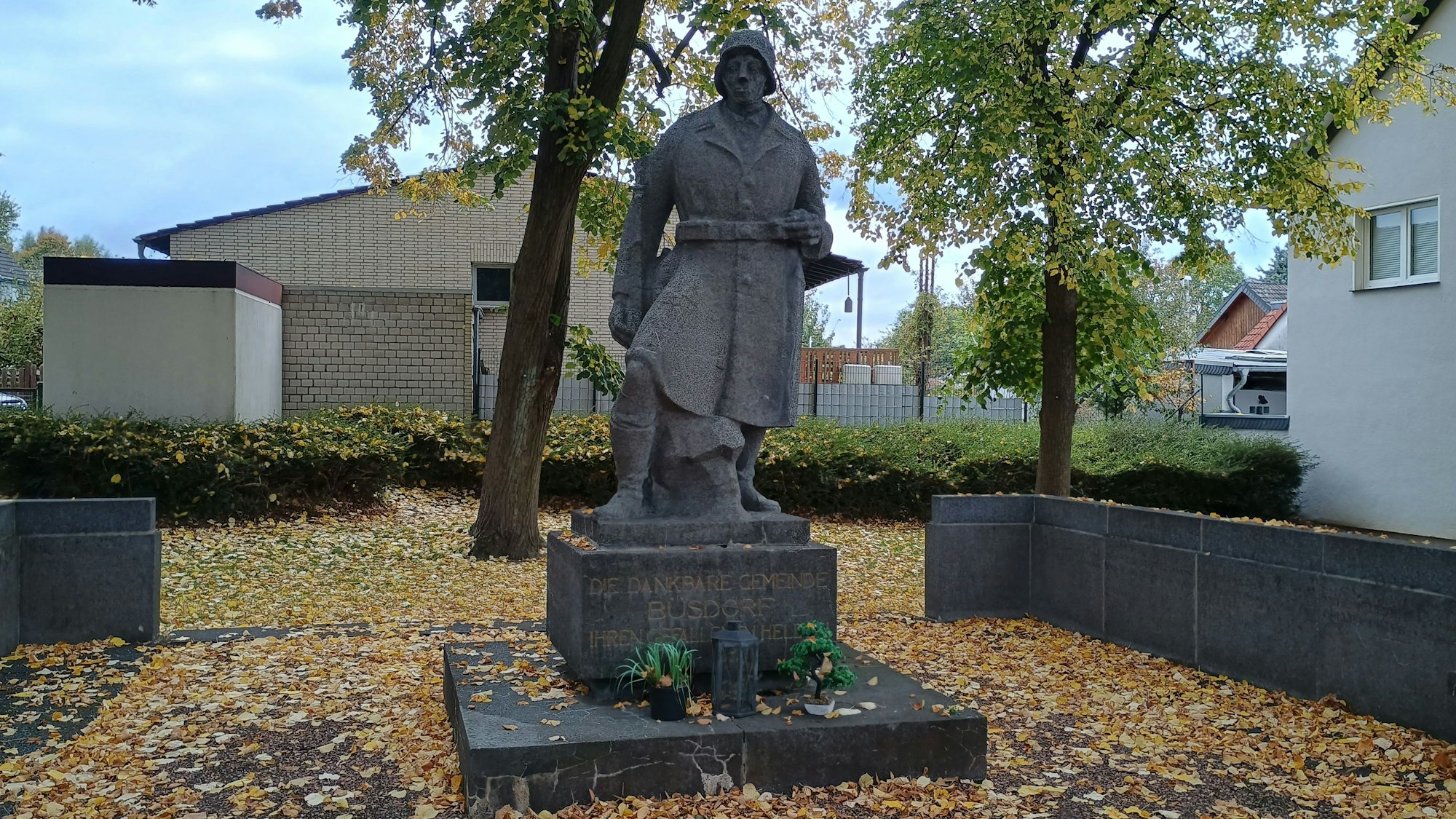 Zu sehen ist ein Steinsoldat mit Helm und Uniform. Auf seinem Podest steht: „Die dankbare Gemeinde Büsdorf ihren gefallenen Helden.“