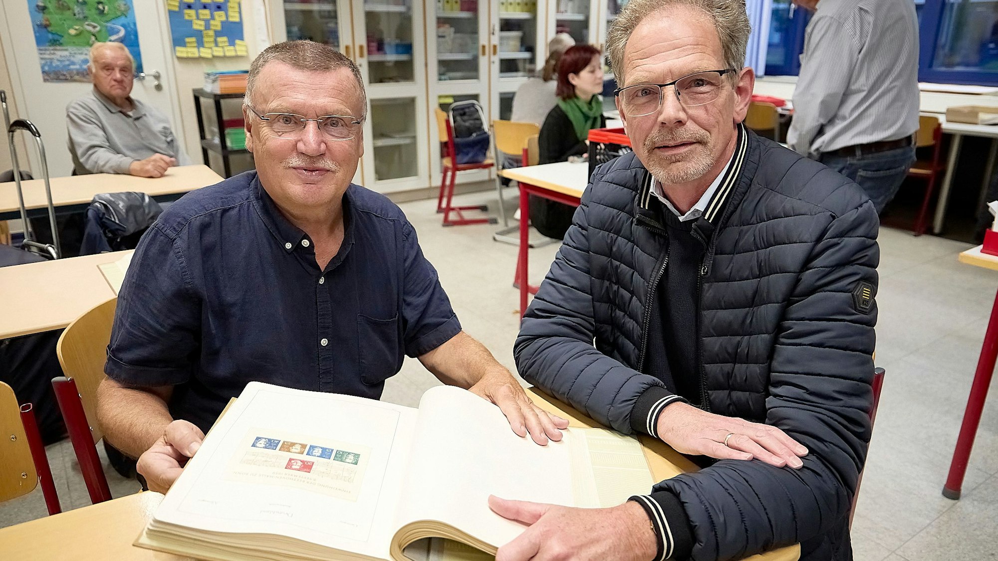 Zwei Männer sitzen an einem Tisch und schauen sich ein Album mit einer Briefmarkensammlung an.