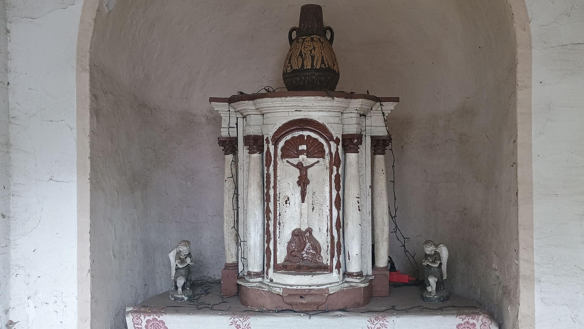 Neben dem hölzernen Altar stehen kleine Figuren. Auf ihm steht eine Vase.