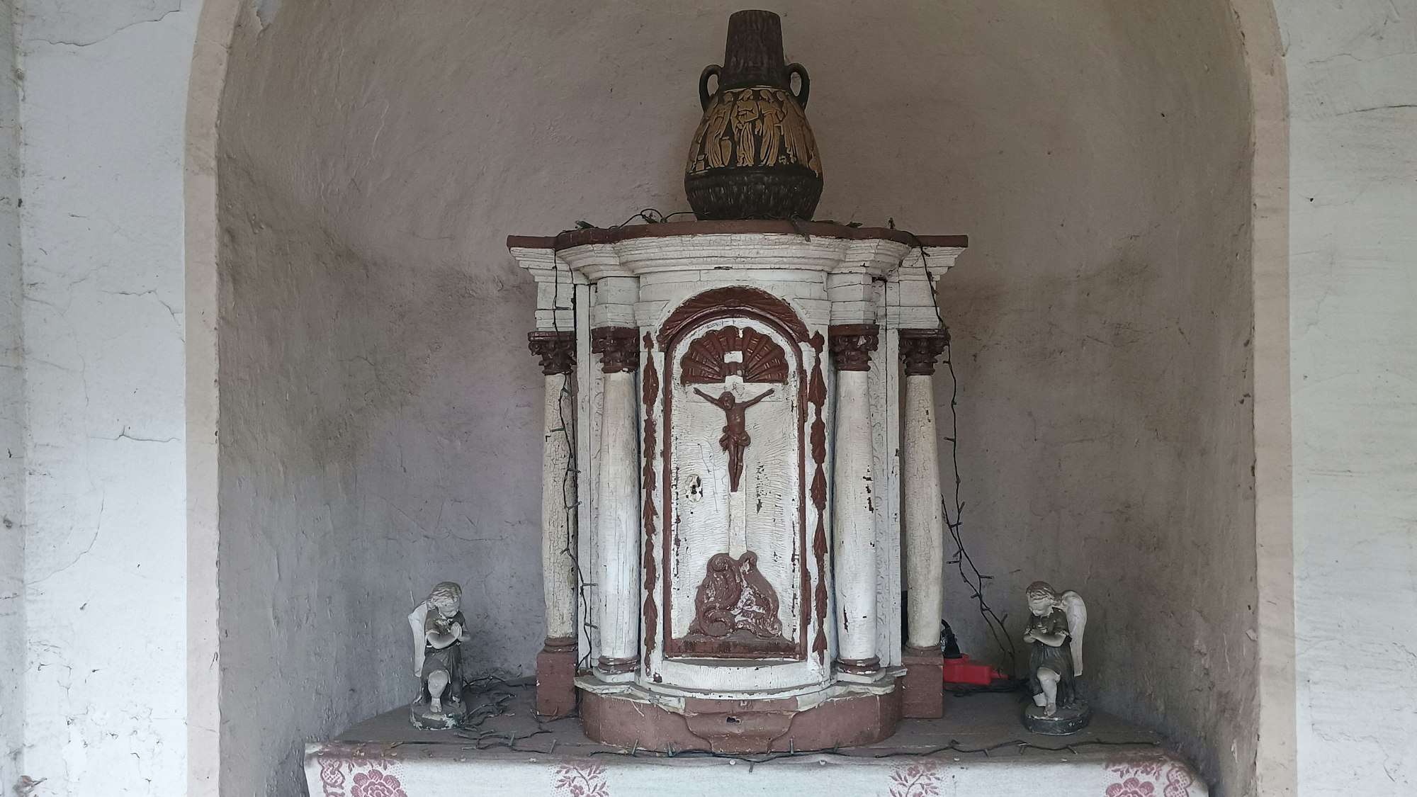 Neben dem hölzernen Altar stehen kleine Figuren. Auf ihm steht eine Vase.