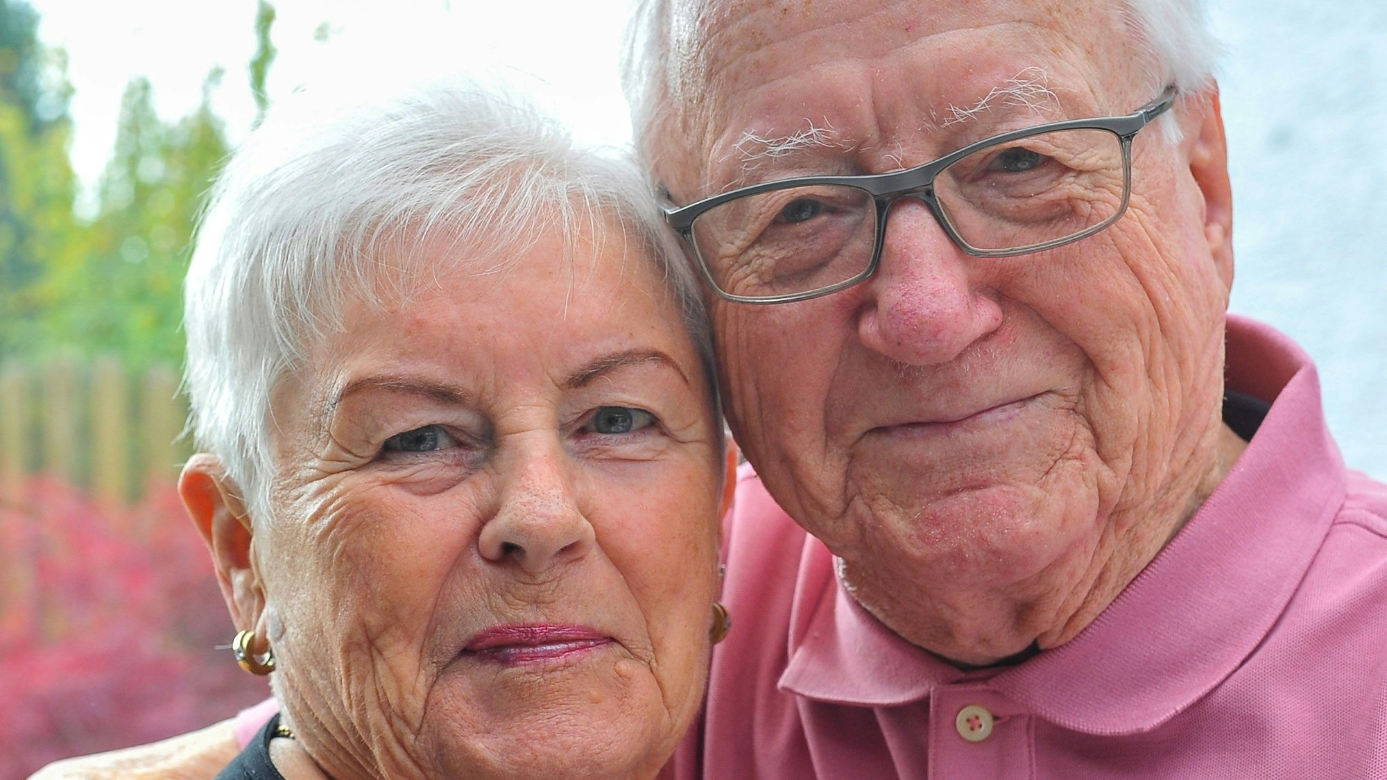 Eine Frau und ein Mann lächeln in die Kamera. Er hat beide Arme um sie gelegt.