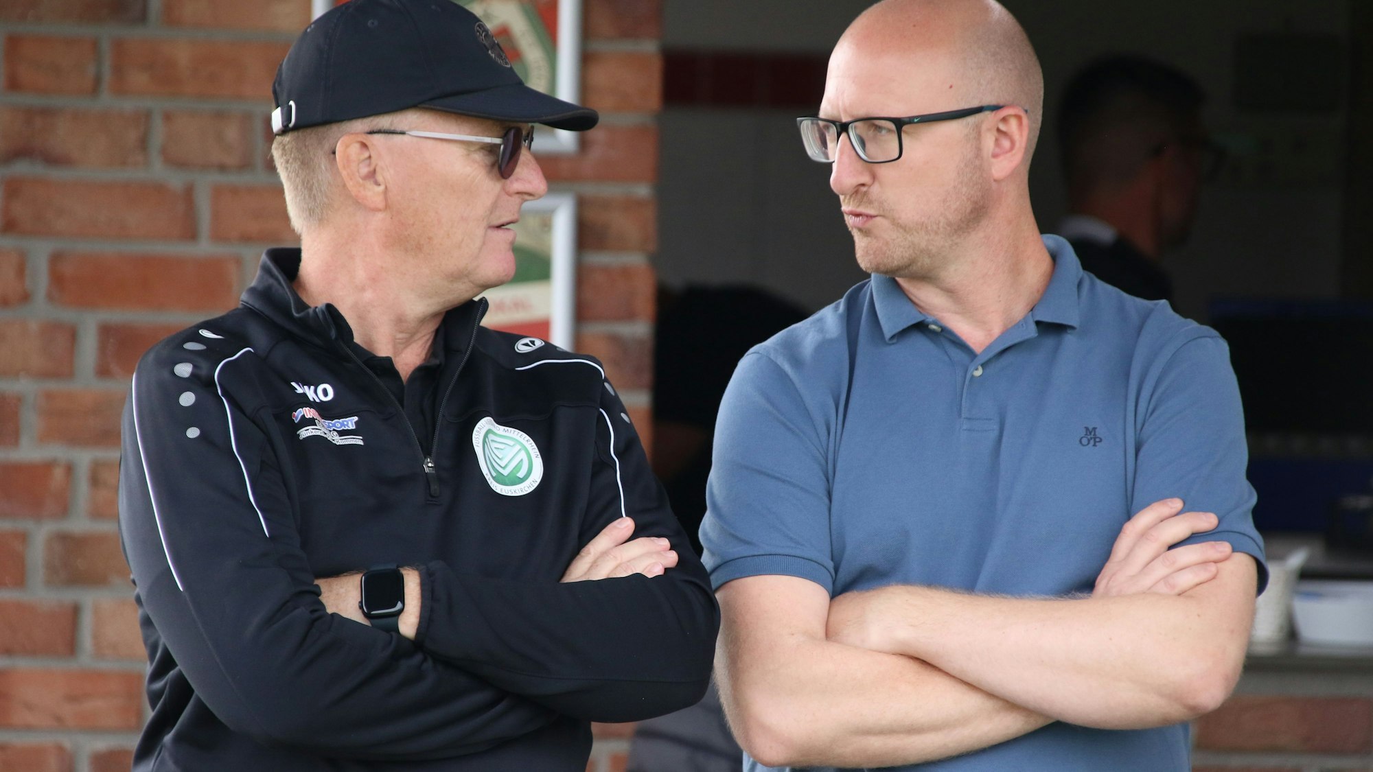 Peter Dierichsweiler mit offiziellem Fußballkreis-Oberteil, mit Kappe und Sonnenbrille unterhält sich mit Ingo Pfennings. Beide schauen sich an.