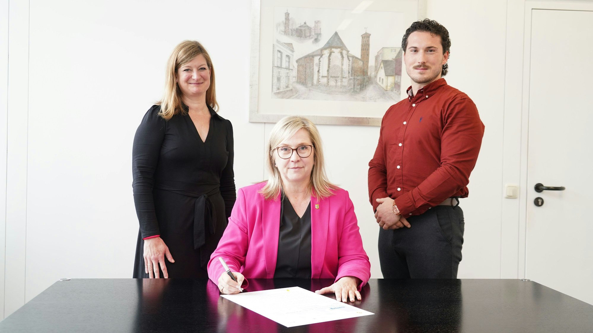Das Foto zeigt zwei Frauen und einen Mann bei der Unterzeichnung eines Dokuments.