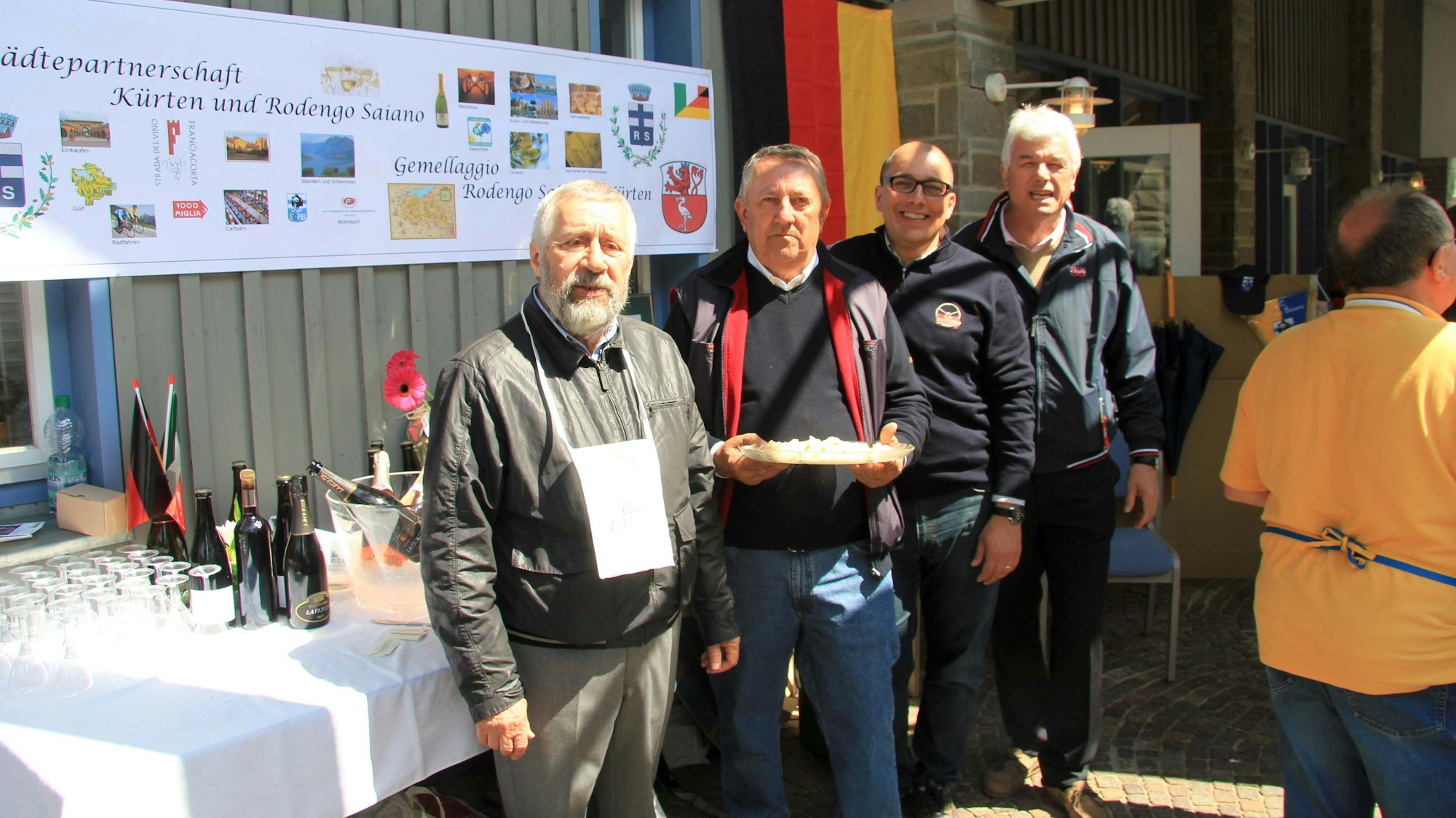 Vier Mitglieder der deutsch-italienischen Städtepartnerschaft Kürten-Rodengo Saiano stehen nebeneinander vor einem Tisch. Einer von ihnen hält etwas zu essen in der Hand.