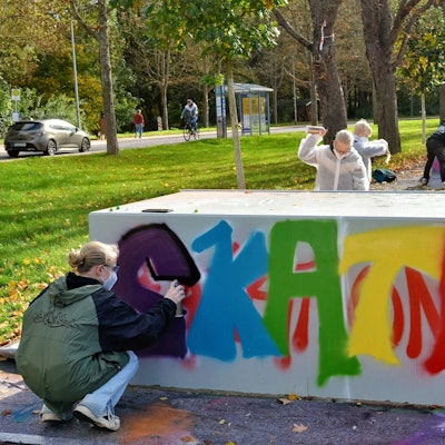 Jugendliche bringen mit Spraydosen Farbe auf die graue Skateanlage. Das Wort „Skate“ ist zu lesen, die Buchstaben in unterschiedlichen Farben.