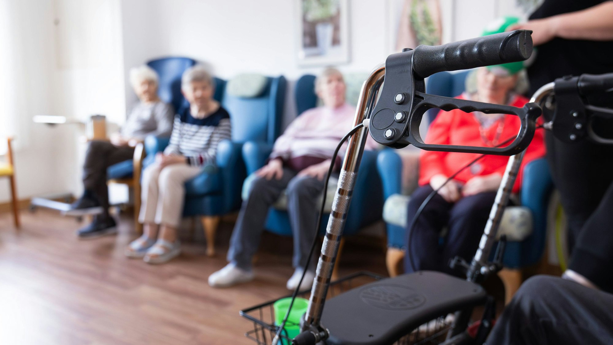 Rollator im Vordergrund, unscharf im Hintergrund sitzende Seniorinnen in einem Aufenthaltsraum eines Altenheims.