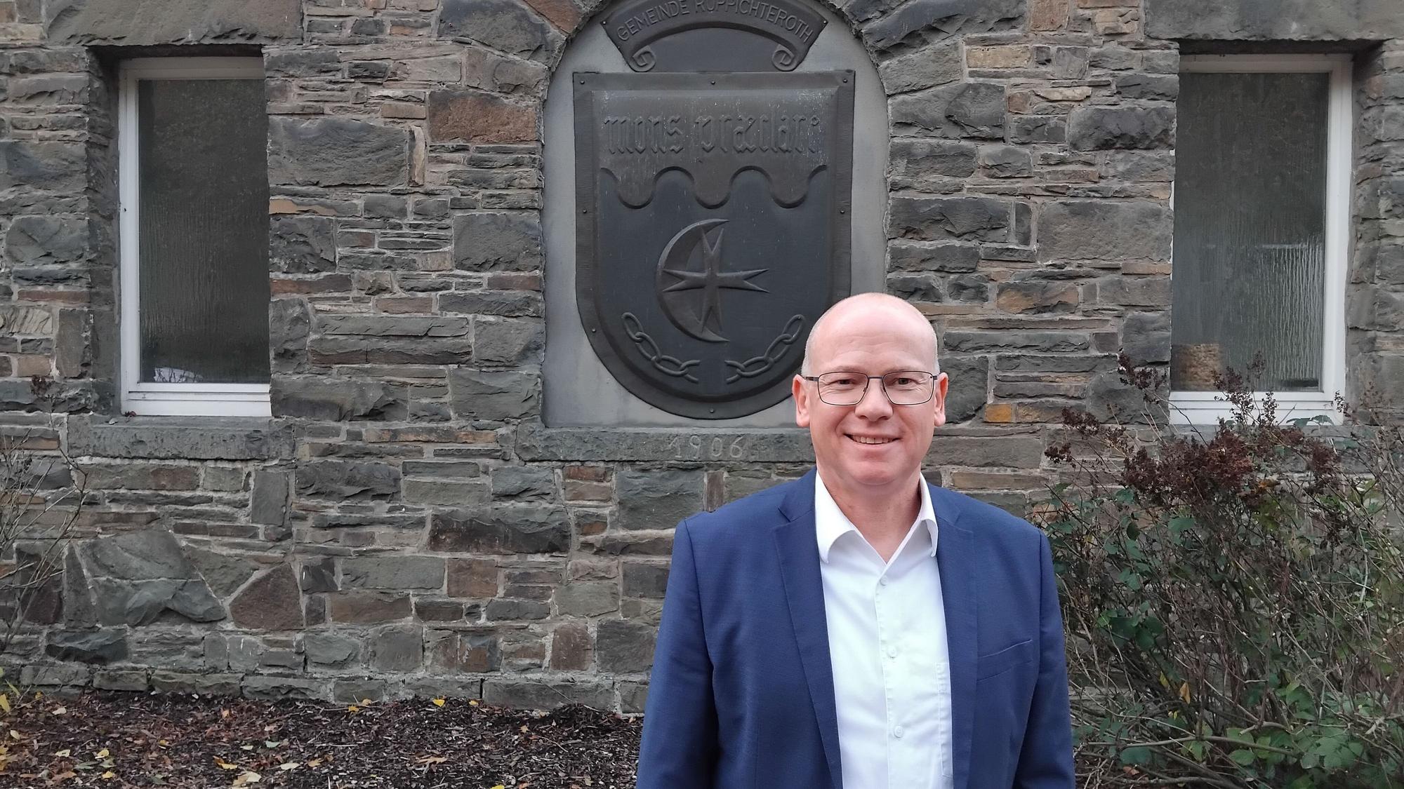 Mario Loskill, Bürgermeister von Ruppichteroth, vor dem Wappen am Rathaus Schönenberg, mit der lateinischen Aufschrift Mons Praeclare (ganz klarer Berg), die für Schönenberg steht.