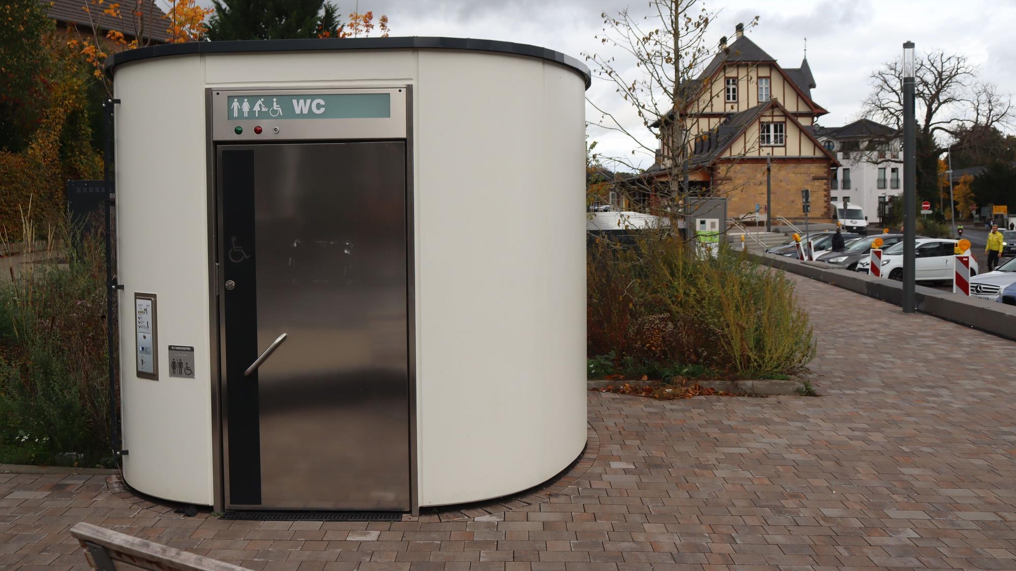 Ein Toilettenhäuschen steht im Vordergrund, im Hintergrund ist das Bahnhofsgebäude zu sehen.