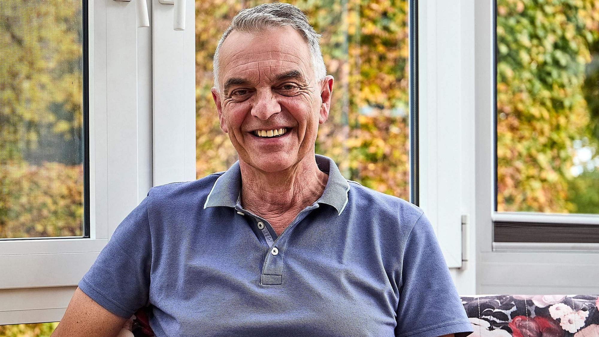 Rudolph Westerburg sitzt in einem blauen T-Shirt vor einem Fenster und lächelt in die Kamera.