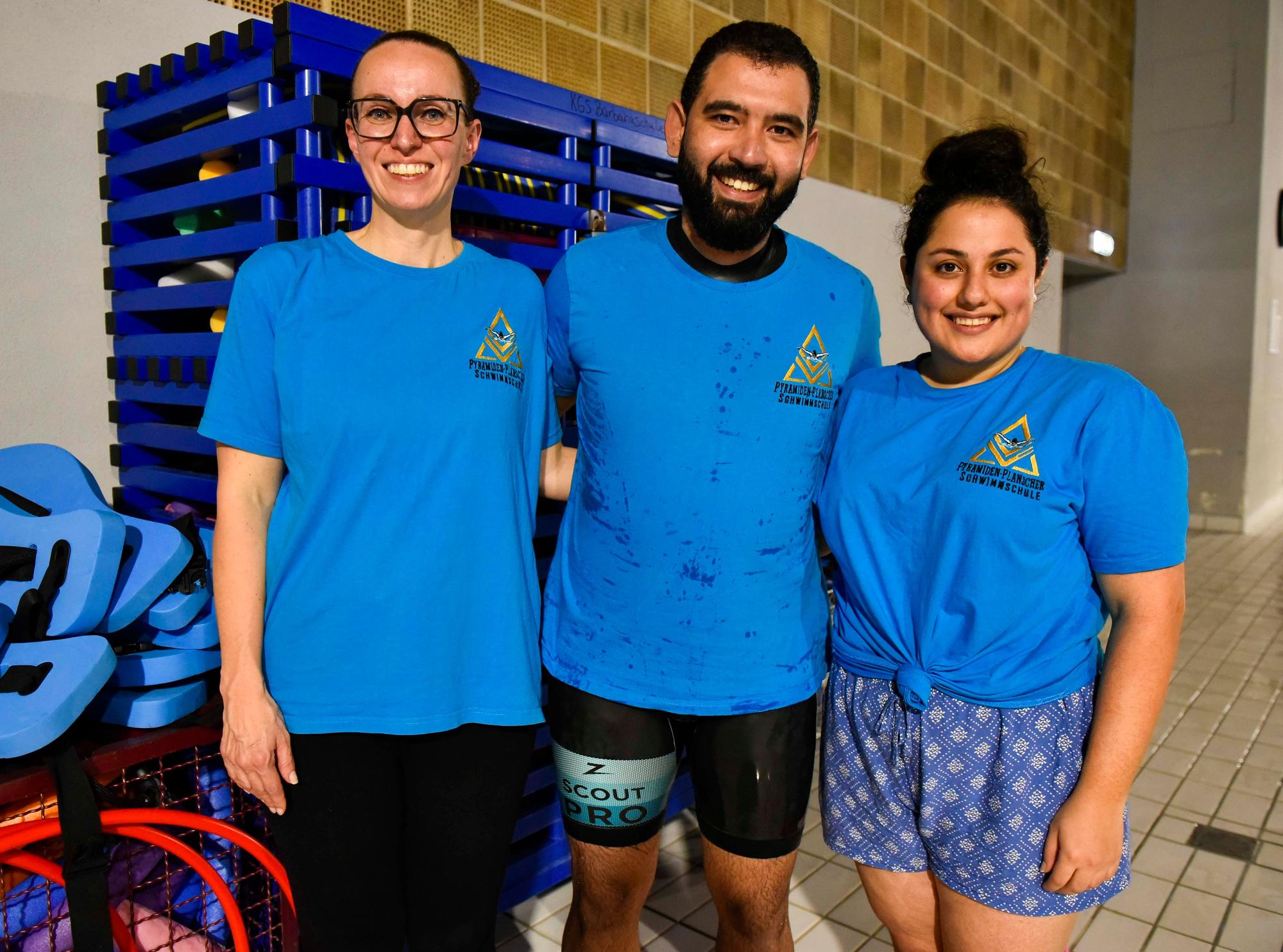 Alle drei tragen blaue T-Shirts. Im Hintergrund das Bad und Schränke mit Utensilien zum Schwimmen.