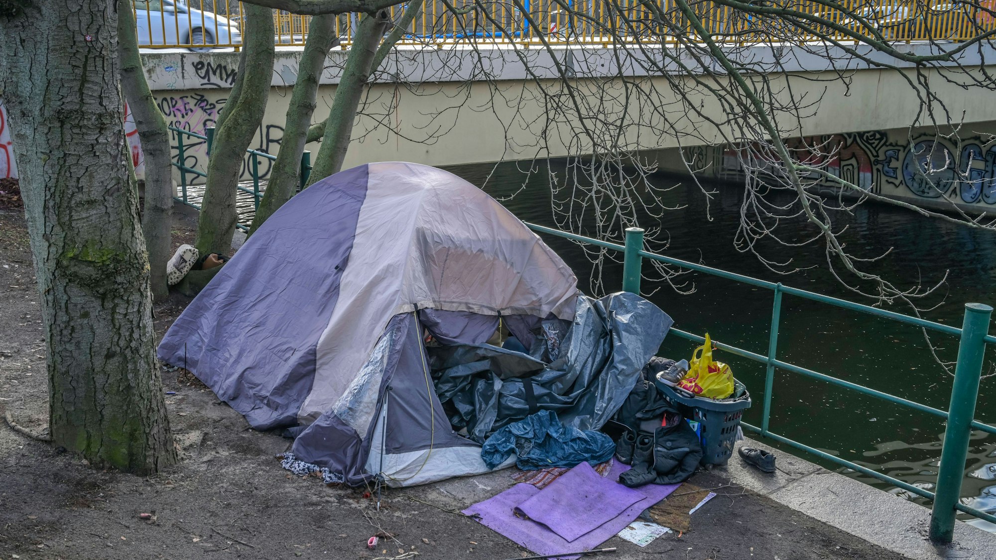 Ein Obdachlosenzelt mit Habseligkeiten am Kanal unter einer städtischen Brücke.