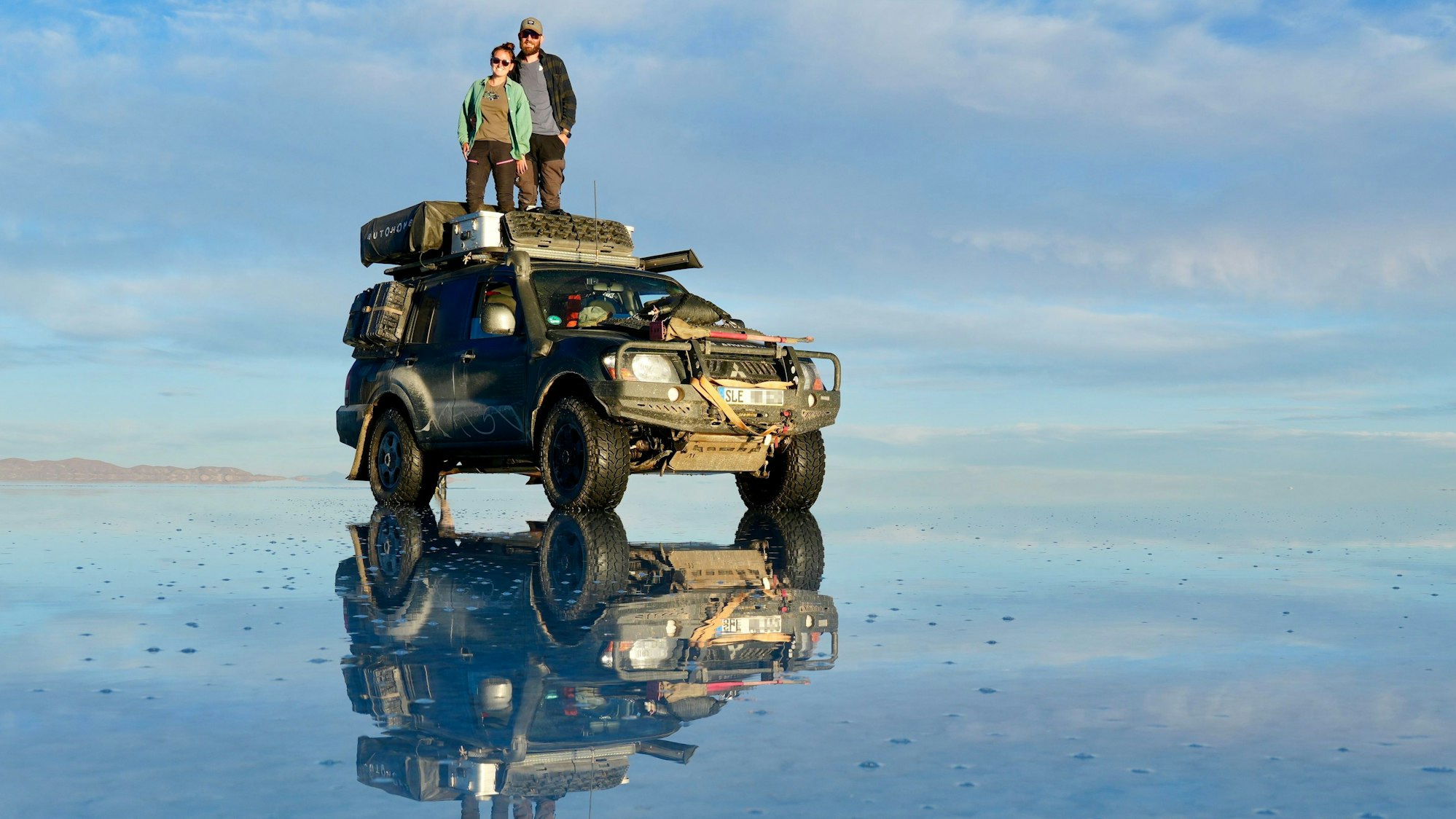 Rebecca und Marcel Schumacher stehen auf dem Dach ihres Mitsubishi Pajero, der sich auf einer Wasserfläche spiegelt.