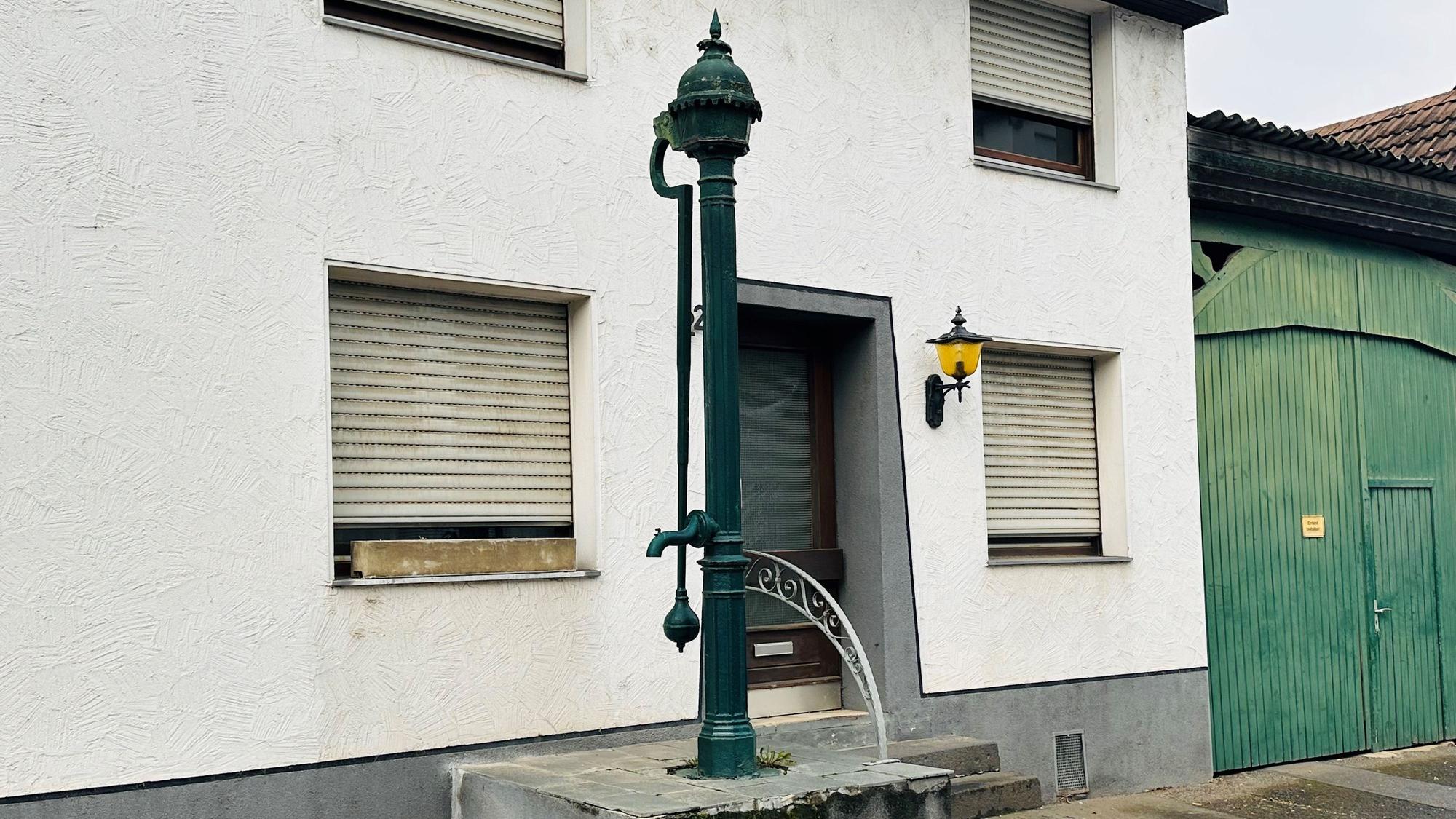 Zu sehen ist eine gusseiserne Pumpe vor einem Haus.