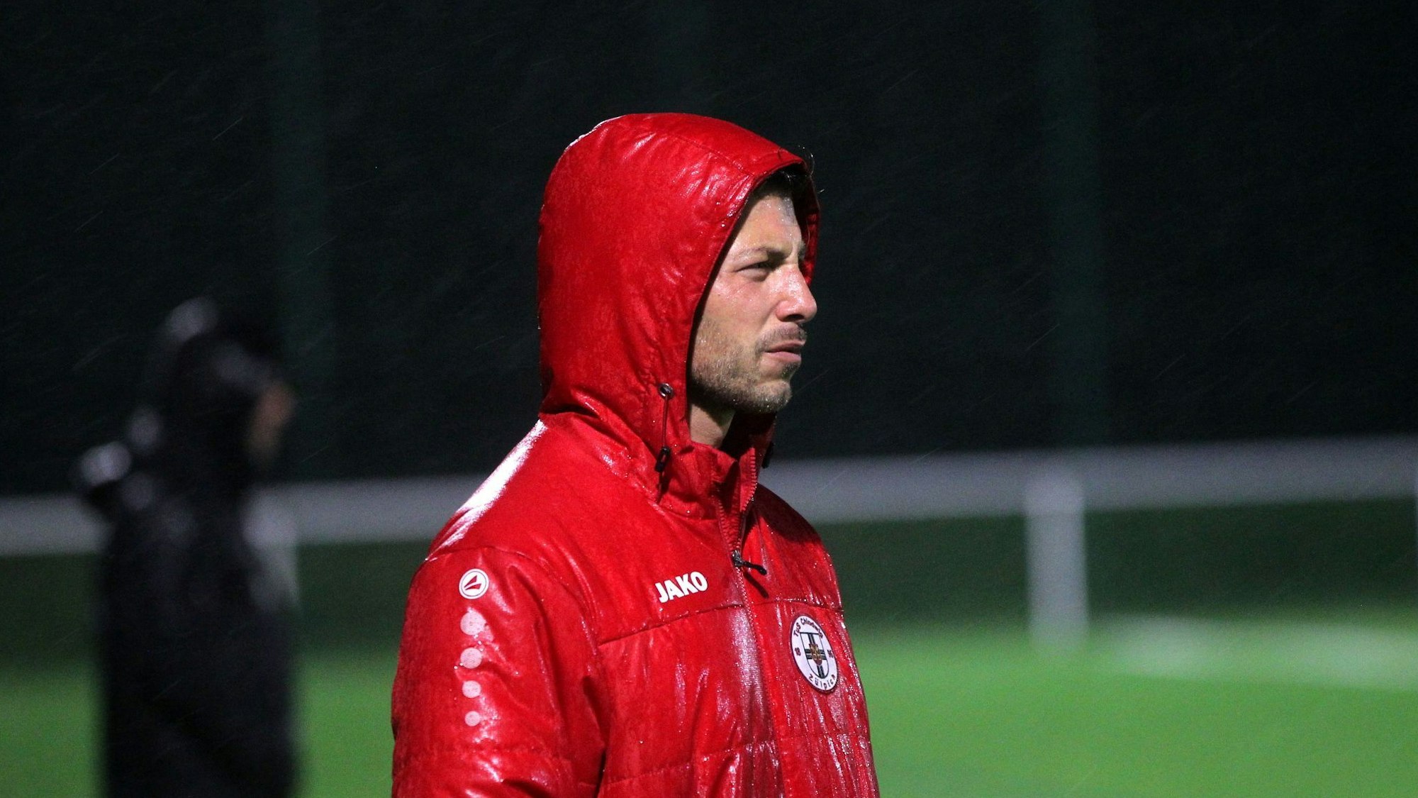 Trainer Marc Altendorf steht in einer nassen roten Jacke am Spielfeld.