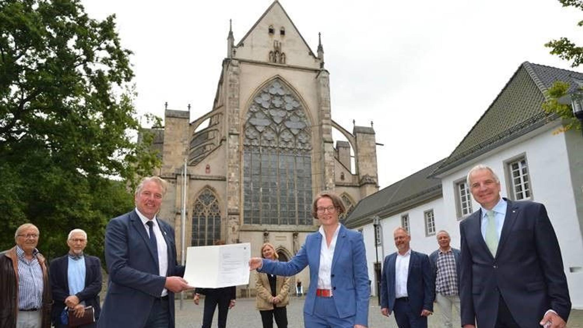 Eine Frau im Hosenanzug überreicht einem Mann im Anzug eine Urkunde. Dahinter der Altenberger Dom.