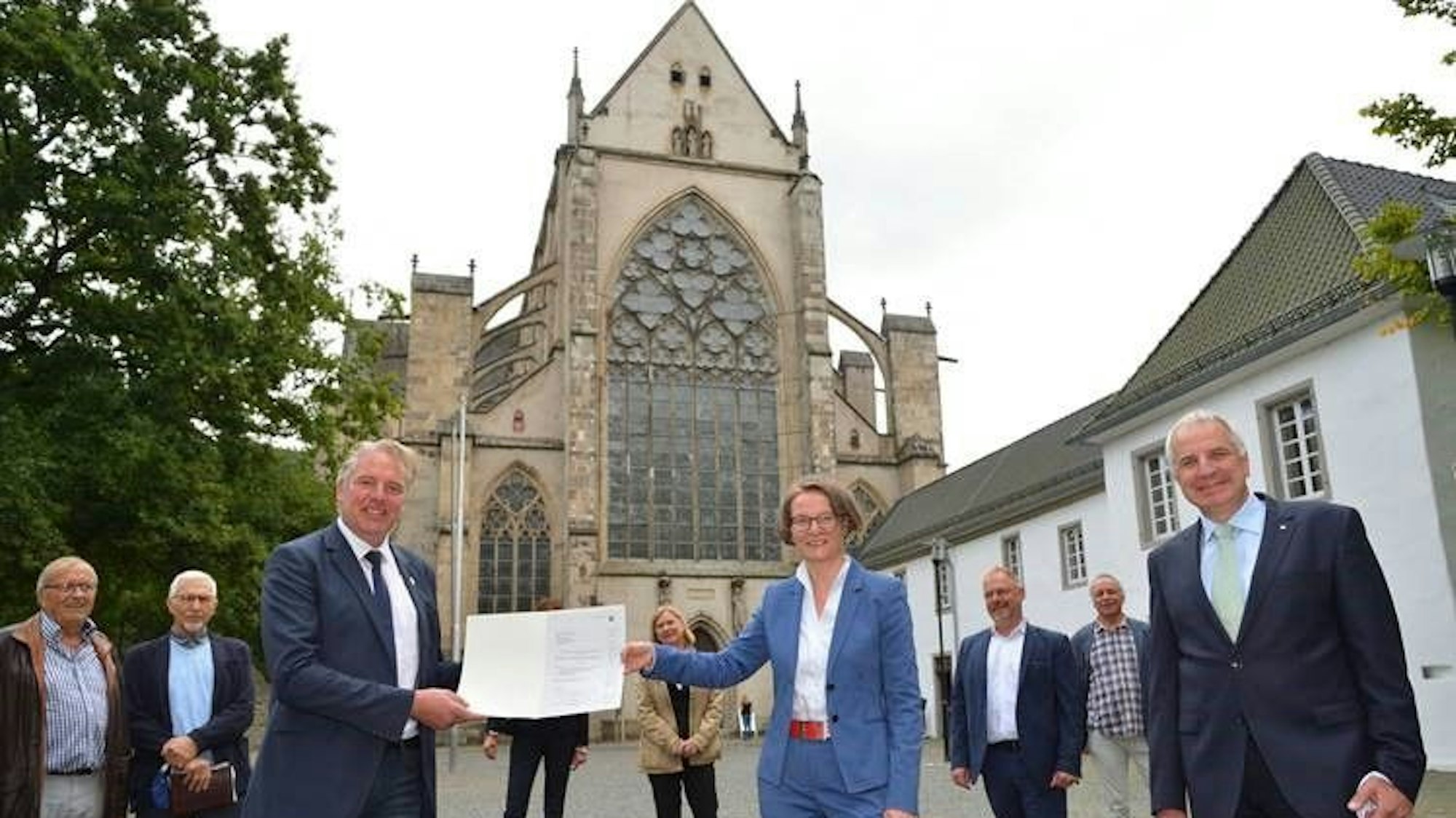 Eine Frau im Hosenanzug überreicht einem Mann im Anzug eine Urkunde. Dahinter der Altenberger Dom.