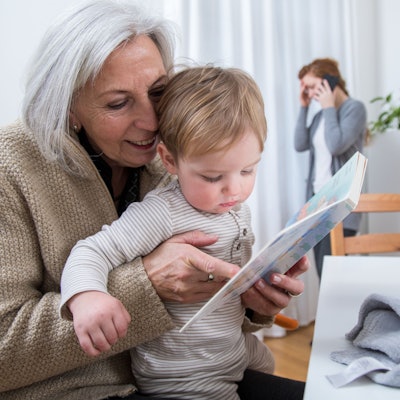 Eine ältere Frau hält in einer Wohnung ihr Enkelkind auf dem Arm, während ihre Tochter im Hintergrund telefoniert