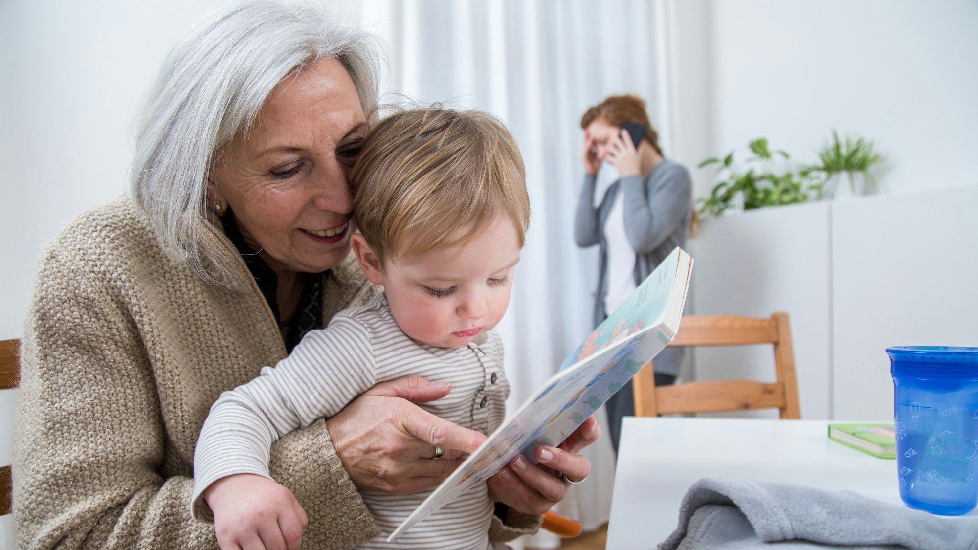 Eine ältere Frau hält in einer Wohnung ihr Enkelkind auf dem Arm, während ihre Tochter im Hintergrund telefoniert