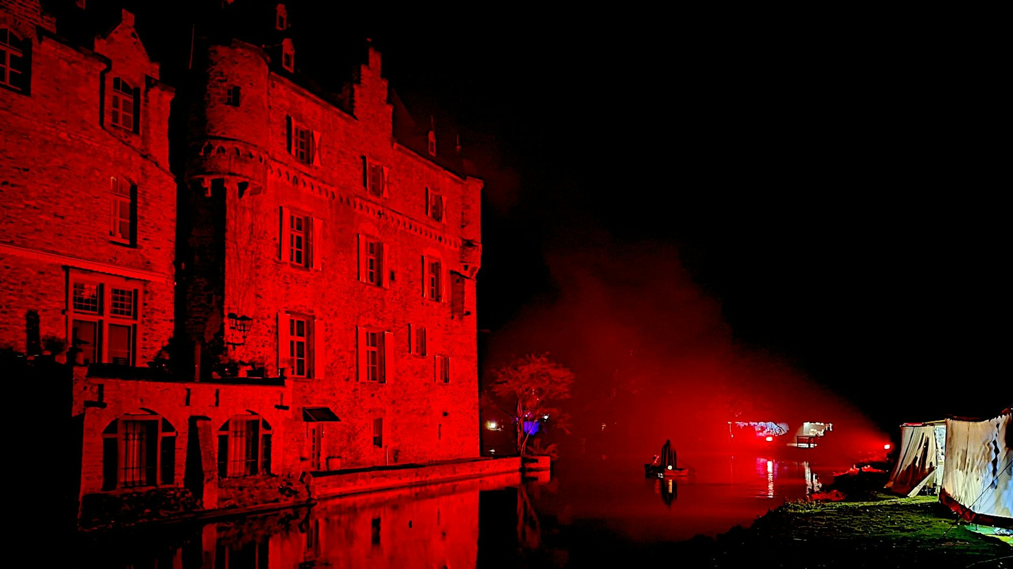 Ein Fährmann ist mit seinem Boot auf dem Burggraben unterwegs. Die Burg Satzvey ist rot angestrahlt.